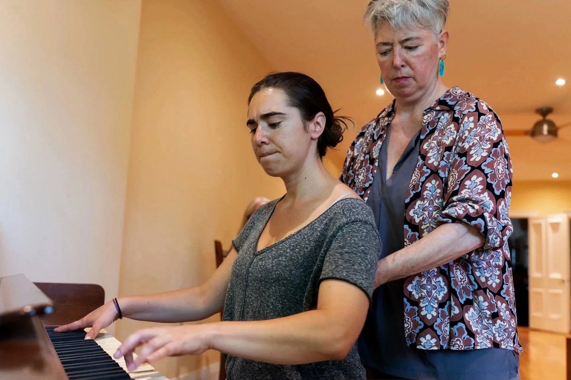 A woman playing the piano while an older woman stands behind her, observing closely.