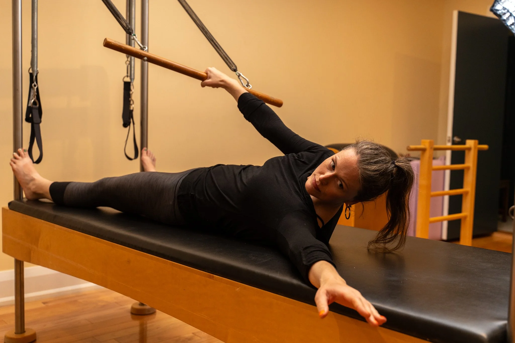 A woman lying on a Pilates reformer machine, holding a wooden bar attached to straps above her head, performing an exercise in a fitness studio.