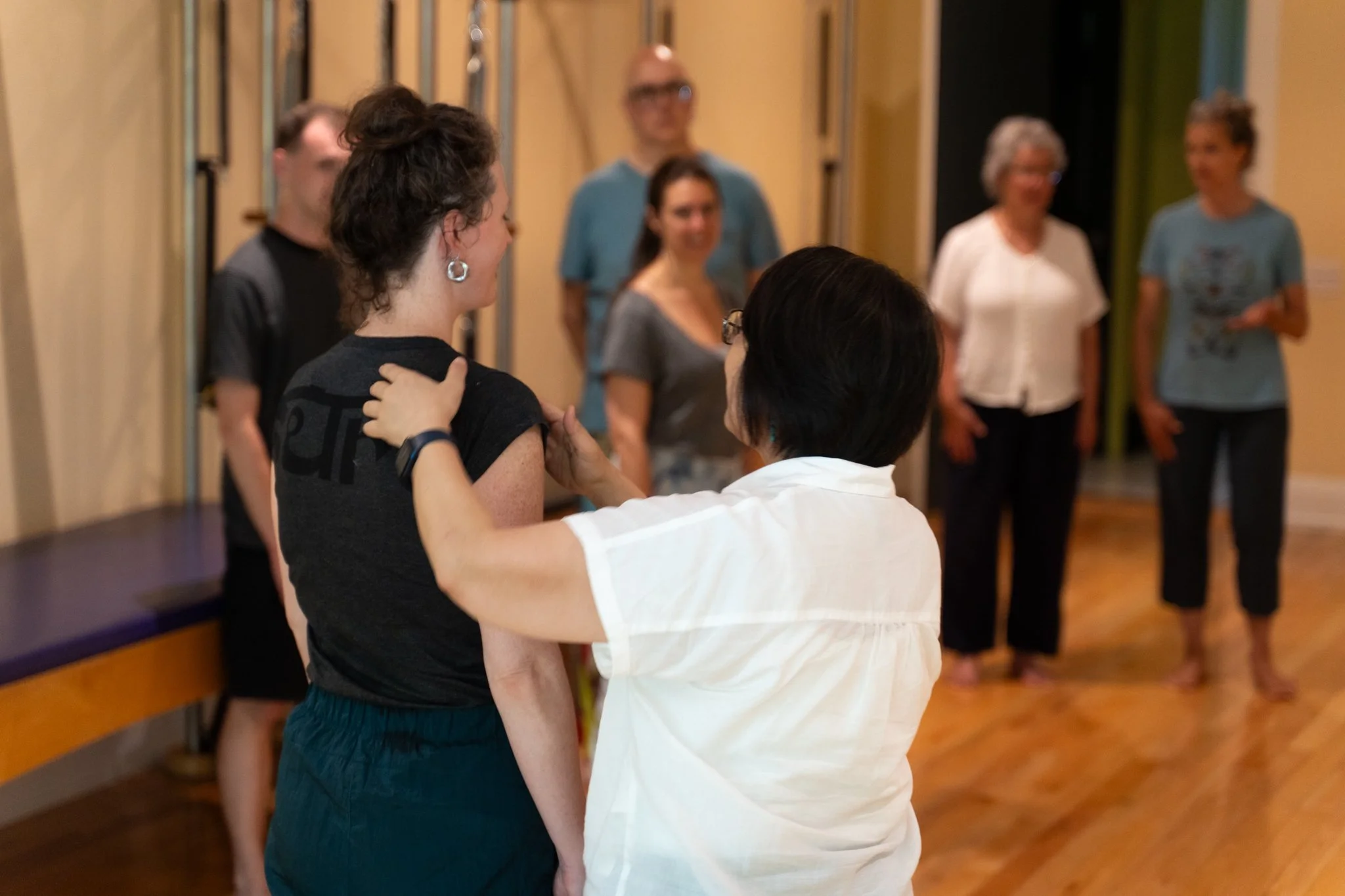 Two women in the foreground are engaged in conversation, with one woman gently touching the other's shoulder. In the background, five people are standing and talking in a casual indoor setting with wooden flooring and beige walls.