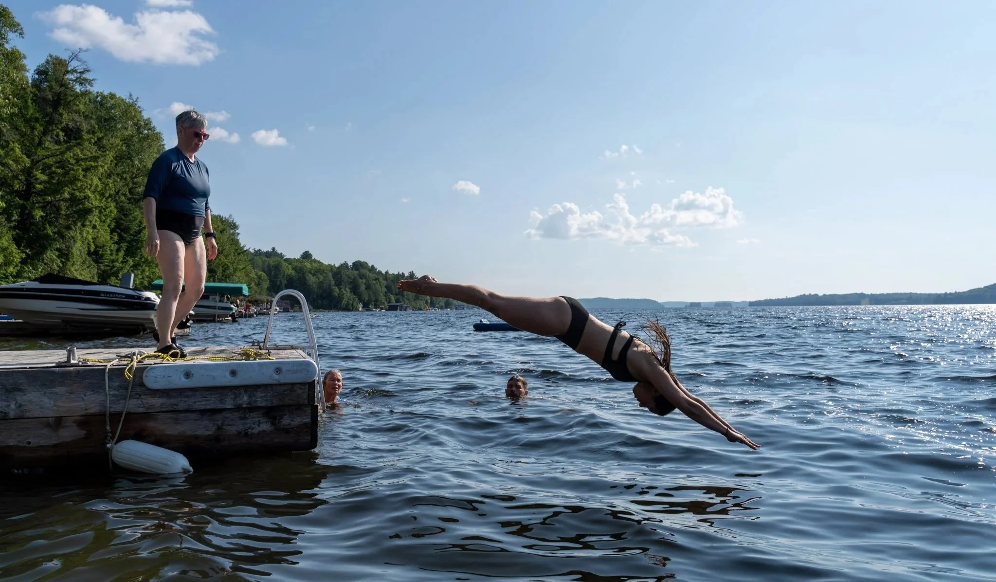 A woman diving into a lake from a dock with two people in the water watching, under a blue sky with some clouds, surrounded by trees and boats.