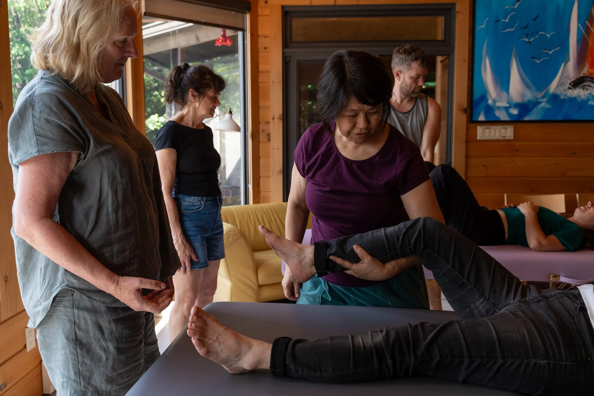 A group of four women and a man participate in a physical therapy or medical training session in a wooden-paneled room with large windows. Two women are standing, observing the session. A woman is assisting a person lying on a table, holding their leg. The person on the table is lying on their back, dressed in black jeans and a green shirt, with their eyes closed.