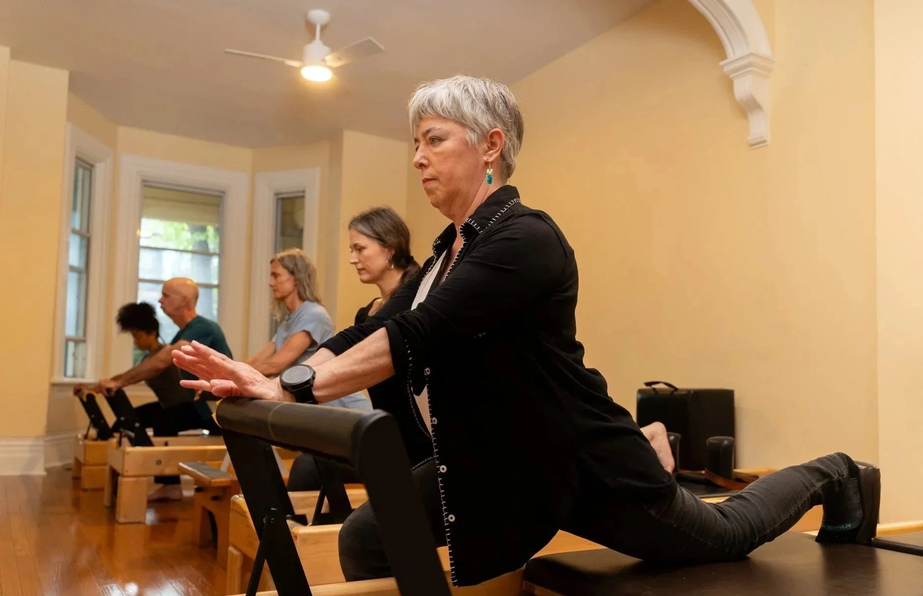 A group of people doing yoga or stretching exercises on benches in a room with large windows, wooden floors, and yellow walls.