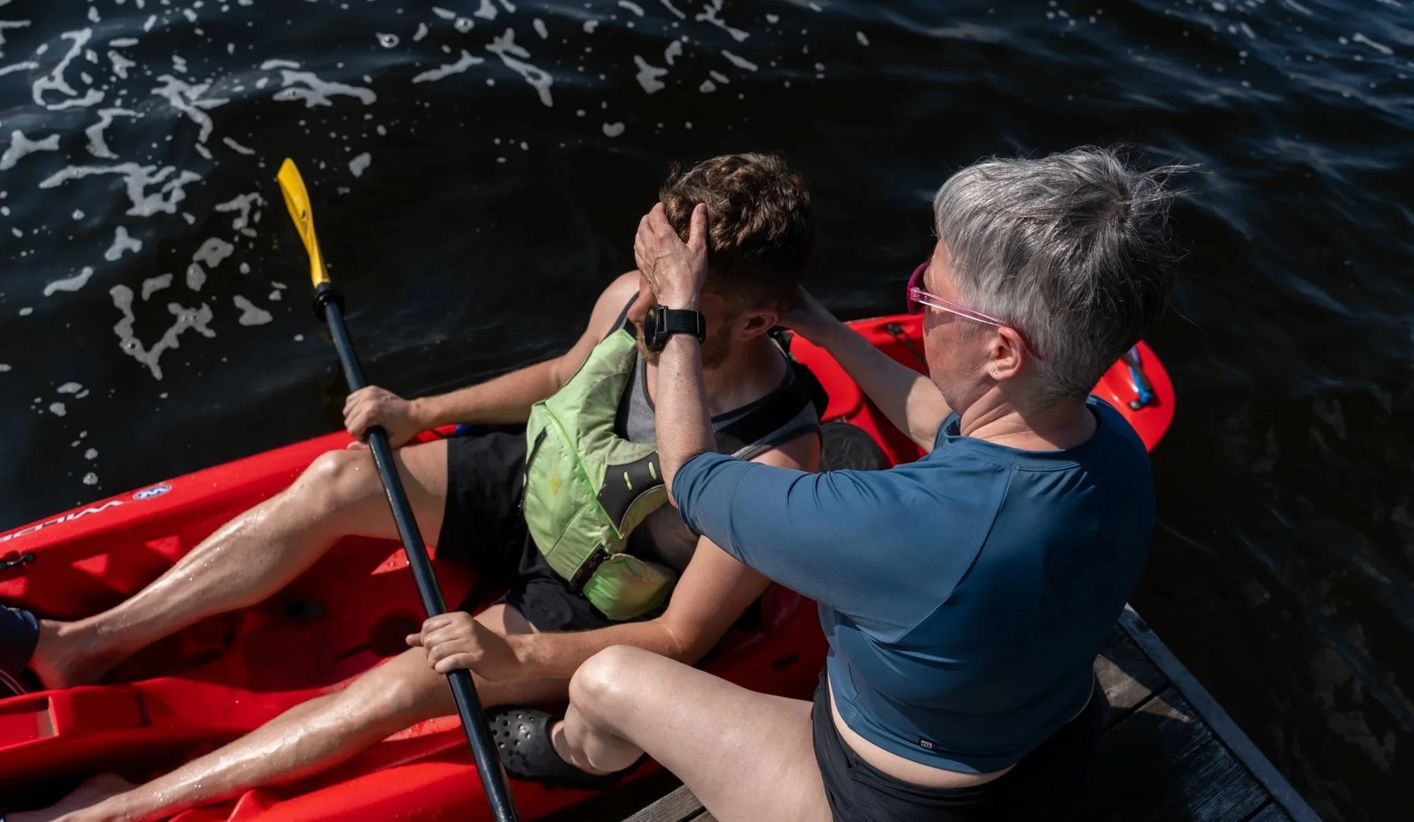 A woman in a blue shirt and glasses comforting a man in a green life vest in a red kayak on dark water, holding his head.