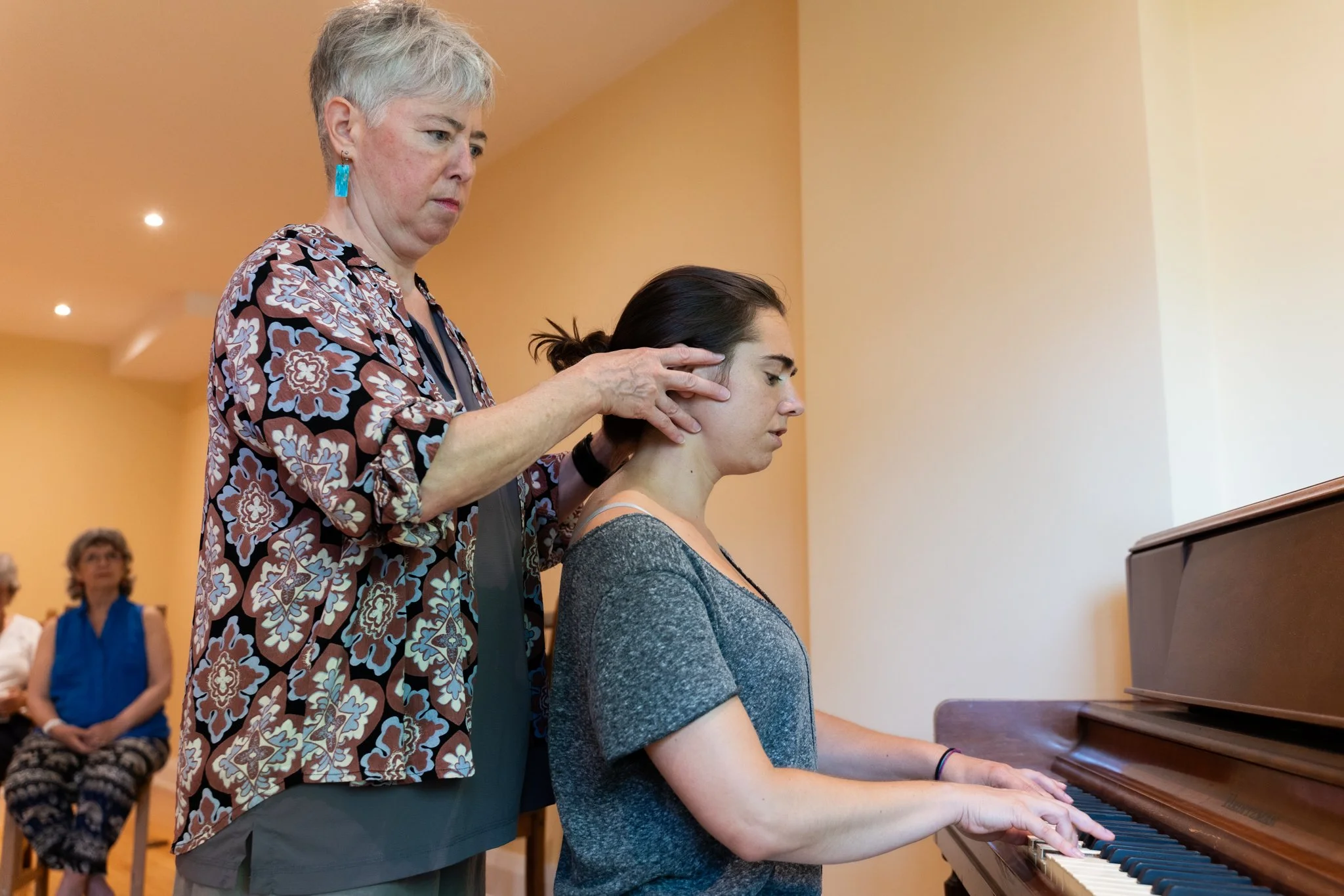 A woman playing the piano while an instructor checks her posture in a room with a few seated spectators.