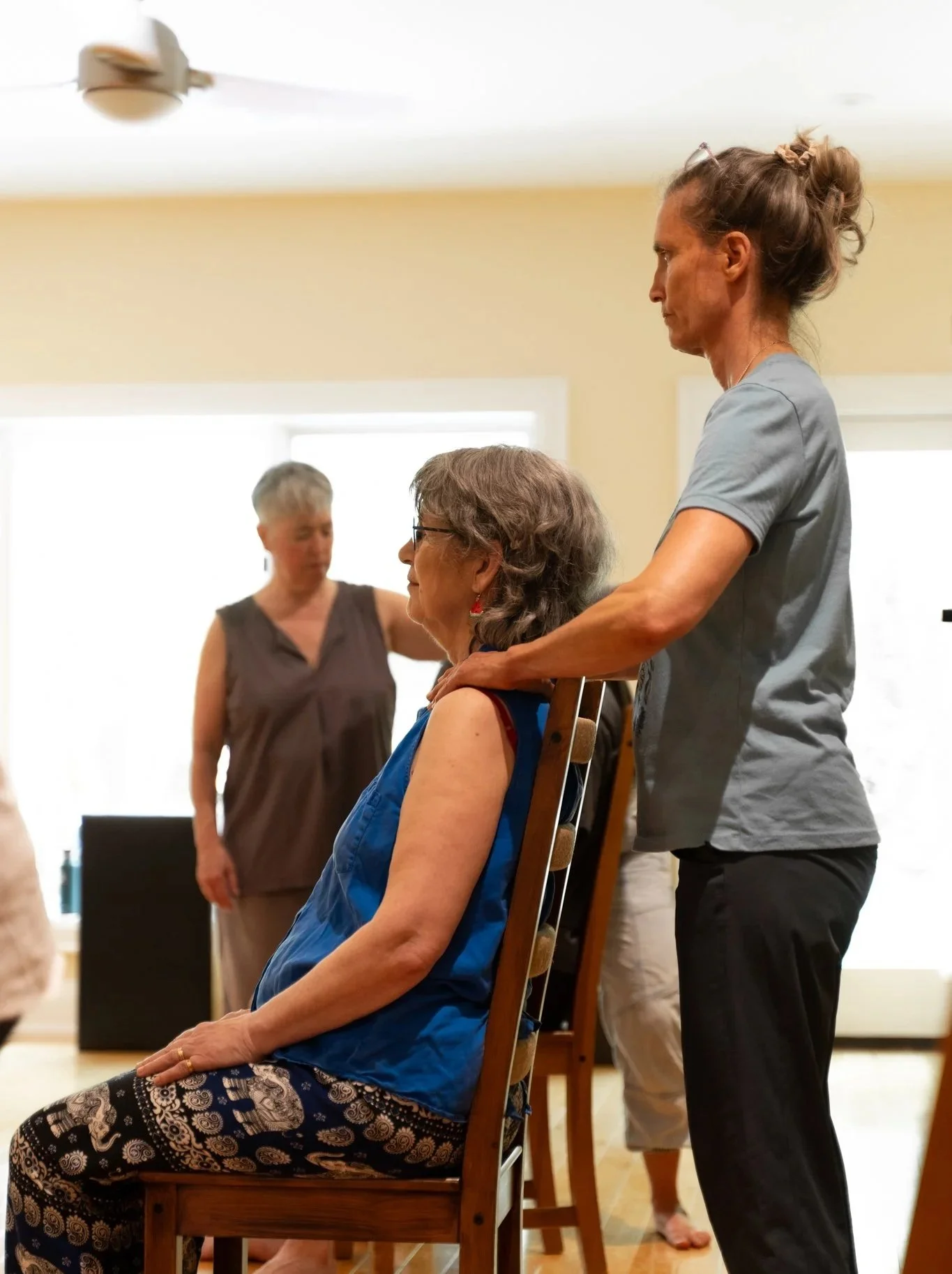 A woman in a blue sleeveless top and patterned pants is seated in a chair, with a caregiver standing behind her, gently supporting her shoulders. Another woman is in the background, standing and observing.