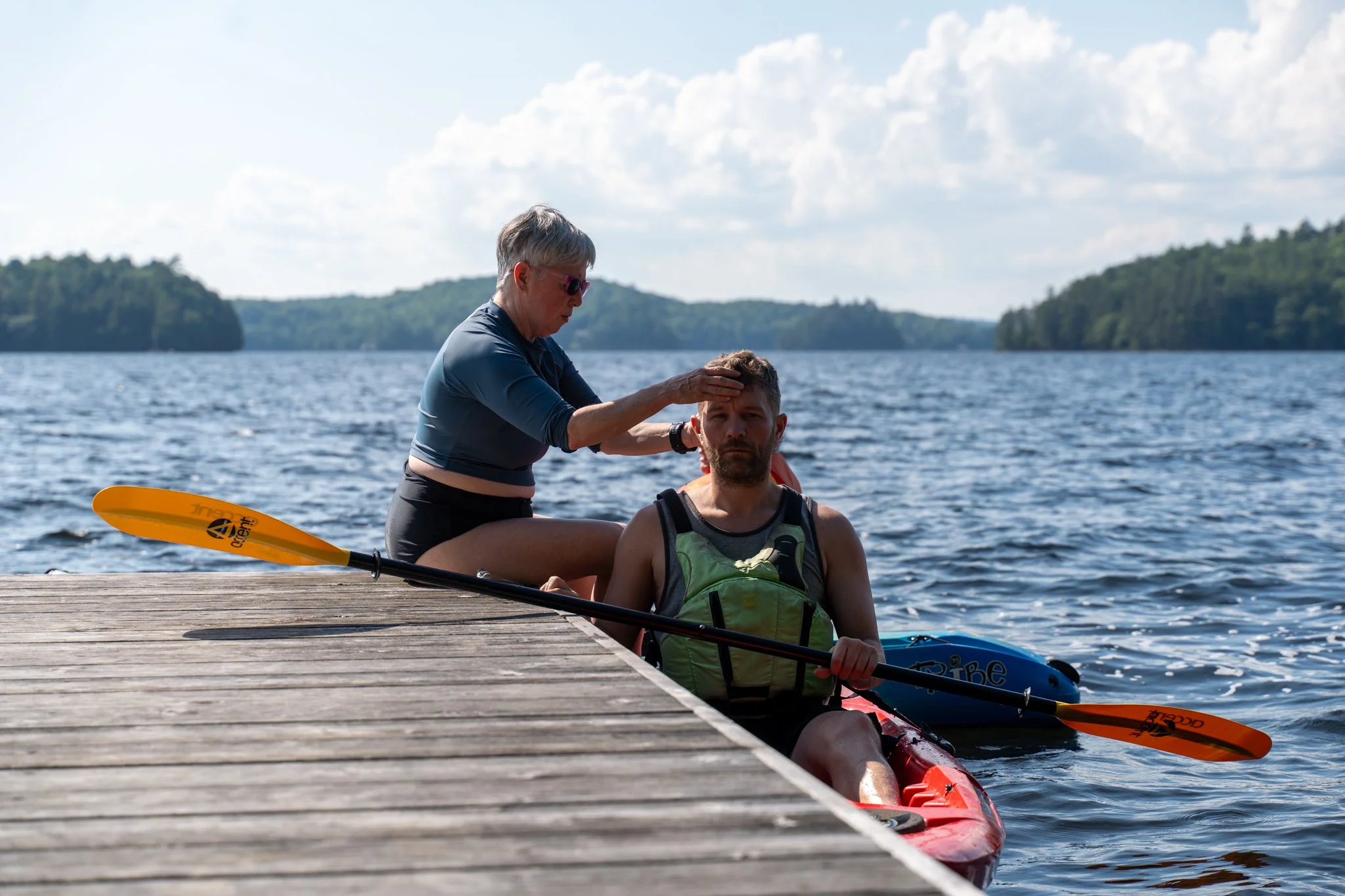 A woman is sitting on a dock, holding a man's shoulder, while the man is sitting in a kayak in the water, holding a paddle. They are on a lake with islands and trees in the background, under partly cloudy skies.