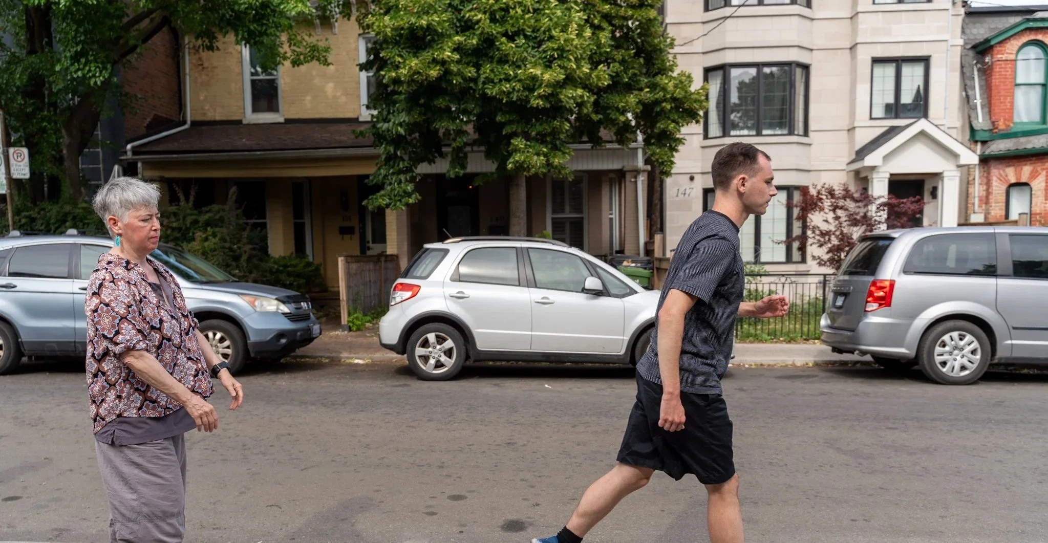 An elderly woman standing on a city street and watching a young man jogging by, with parked cars and residential buildings in the background.