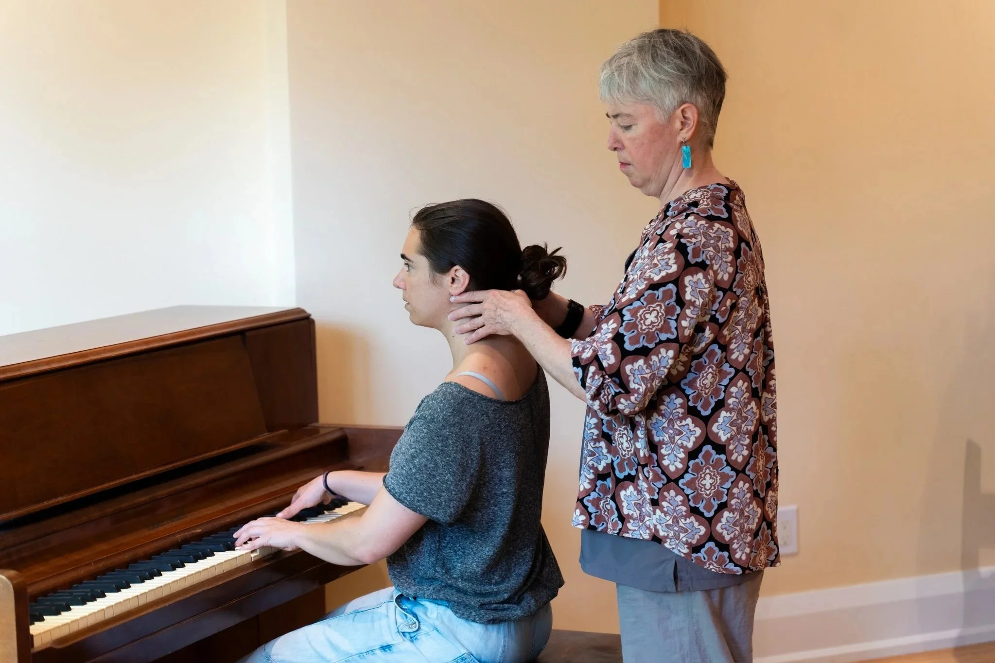 An older woman with short gray hair giving a neck massage to a young woman playing the piano in a room with plain beige walls.