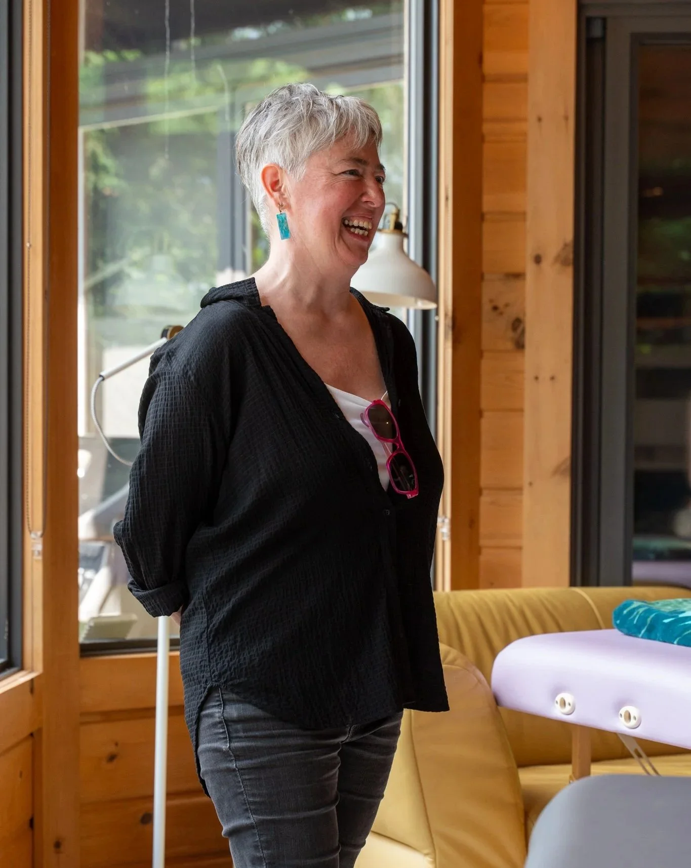 A woman with short gray hair, earrings, and sunglasses hanging from her shirt, smiling inside a wooden cabin with large windows and a yellow couch.