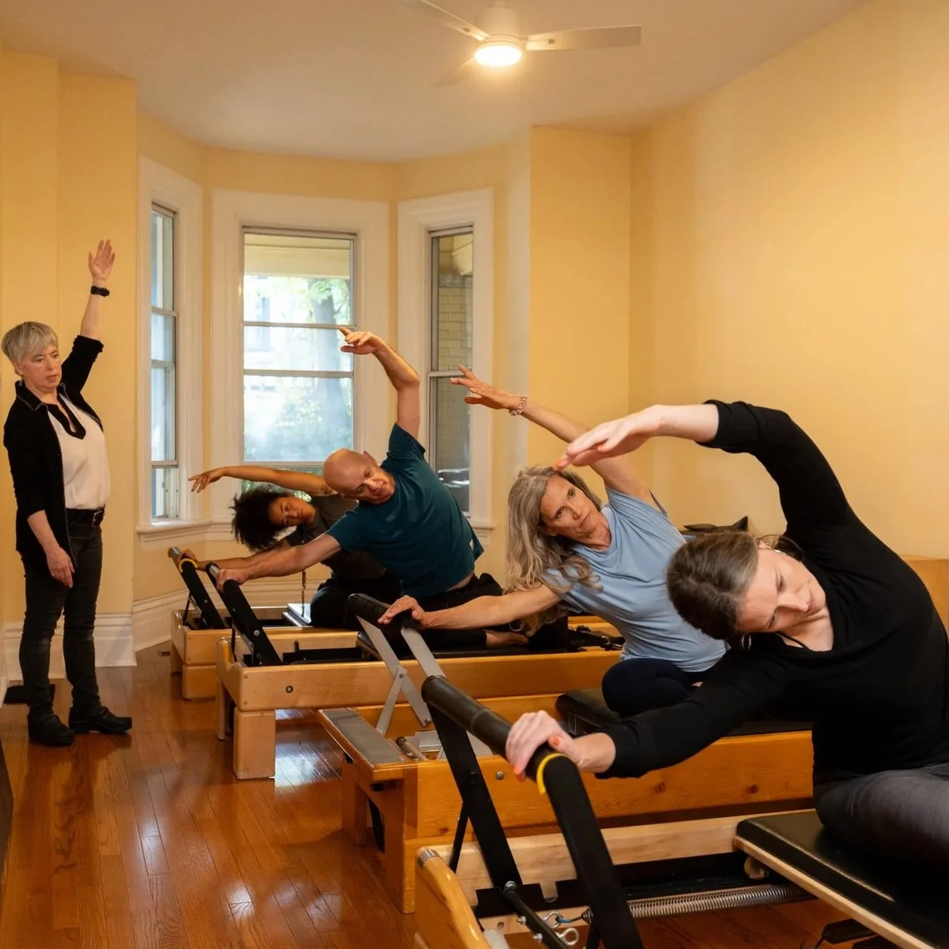 People participating in a Pilates or yoga class in a well-lit room with large windows, using reformer machines to stretch and exercise.