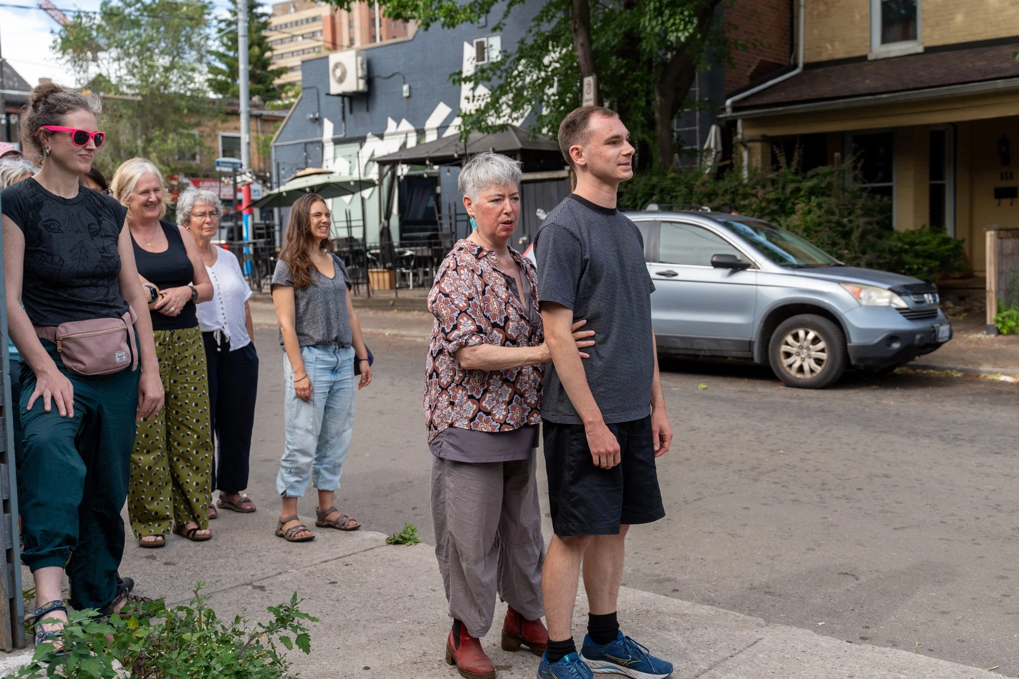 A group of people standing in a line on a sidewalk in an urban neighborhood, with houses and parked cars behind them. The woman in front is holding a young man by the arm, and the group appears to be waiting or observing something.