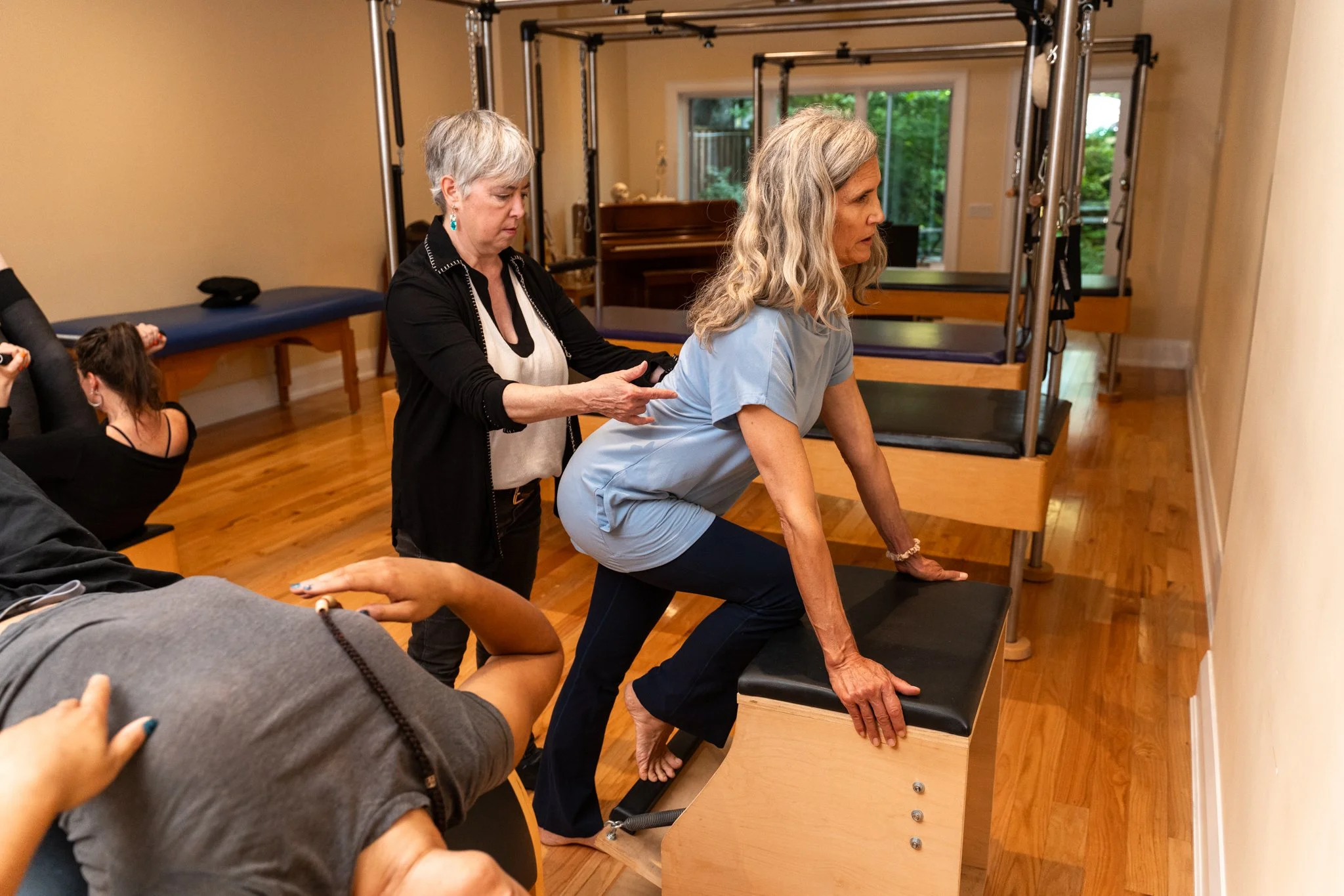 A woman in a light blue top and dark pants is performing a physical therapy exercise by ascending a modified step platform with her hands on it for support, while a therapist with white hair and a black cardigan assists her. Others are present in the background in a room with wooden floors and fitness equipment.