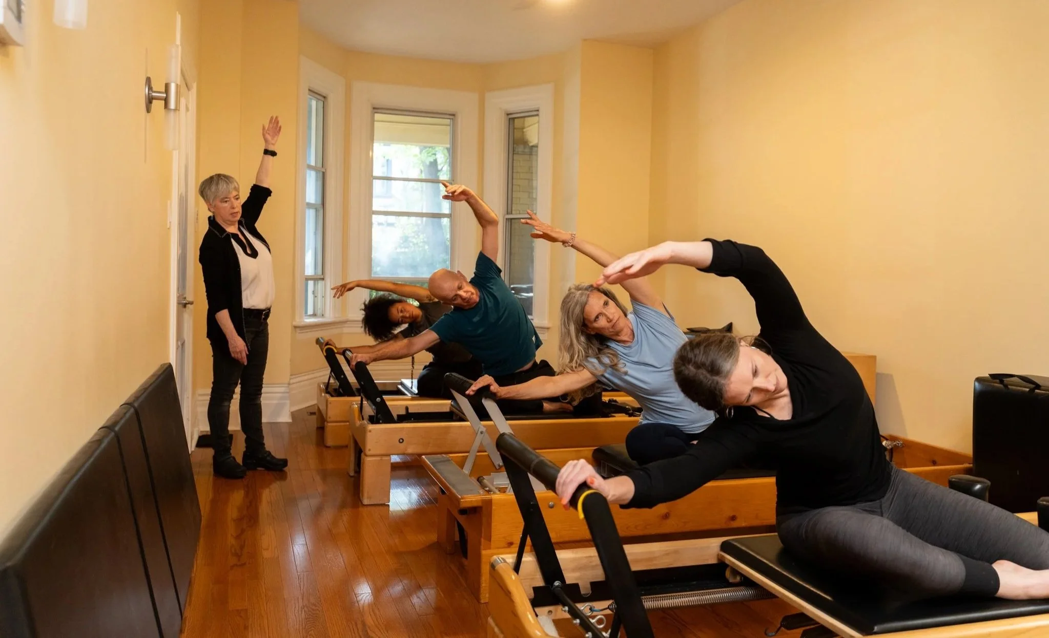 Four women and one instructor participating in a Pilates class on reformer machines in a bright, yellow-walled room with hardwood floors.