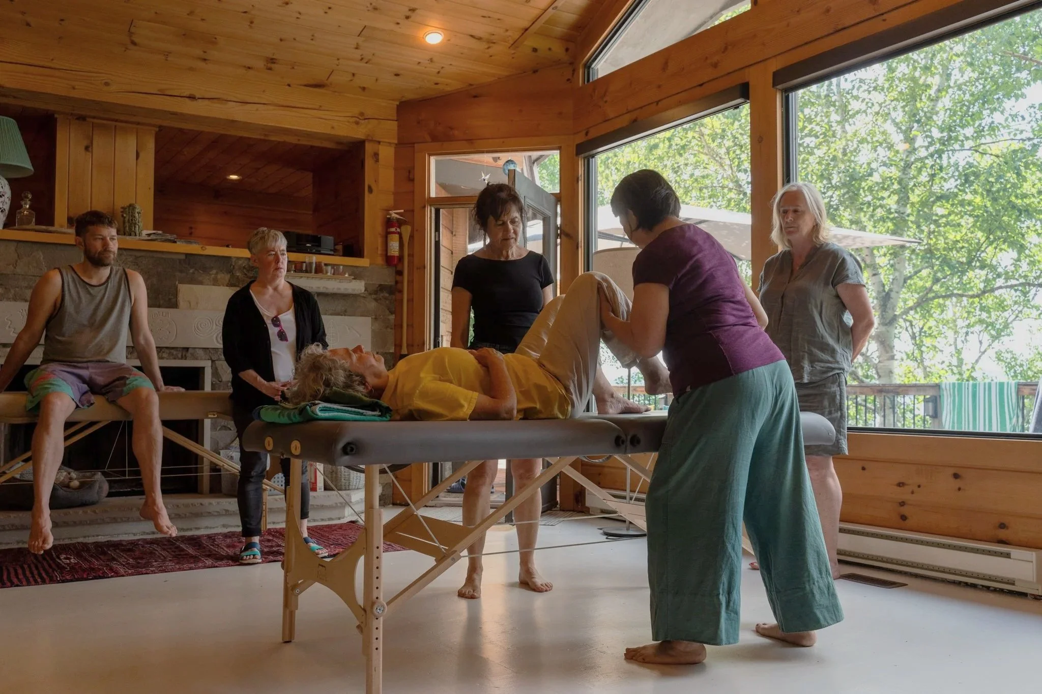 A group of six people in a wooden room, with one woman lying on a massage table, a caregiver performing a procedure, and others observing, with large windows showing trees outside.