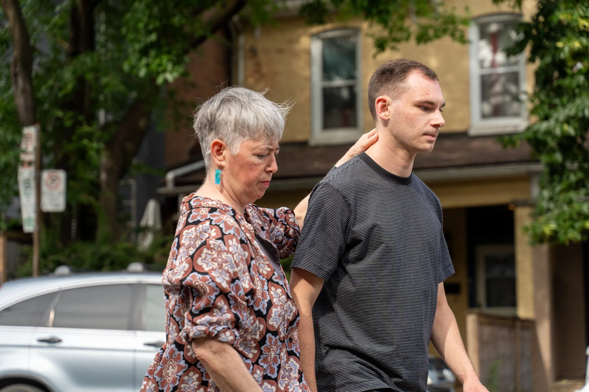 An elderly woman with short gray hair and blue earrings, wearing a patterned blouse, places a hand on the shoulder of a young man with short brown hair, dressed in a black striped T-shirt, walking outdoors in a residential area with trees and houses in the background.