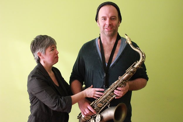 A woman adjusting a man holding a saxophone against a light green background.