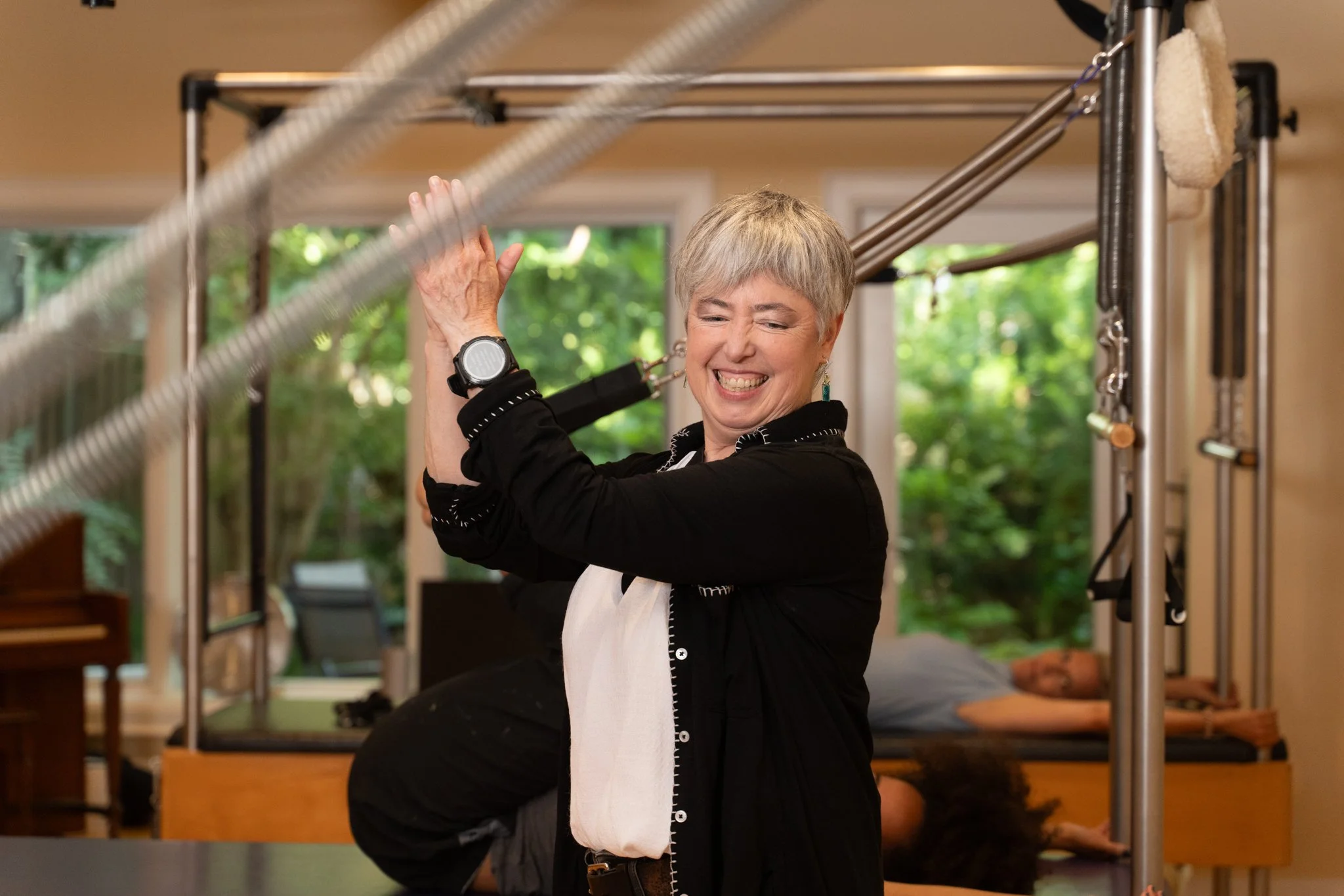 A woman with short gray hair smiling and stretching her arms in a therapy or exercise session, with a person lying on a therapy table in the background and green trees visible through the window.
