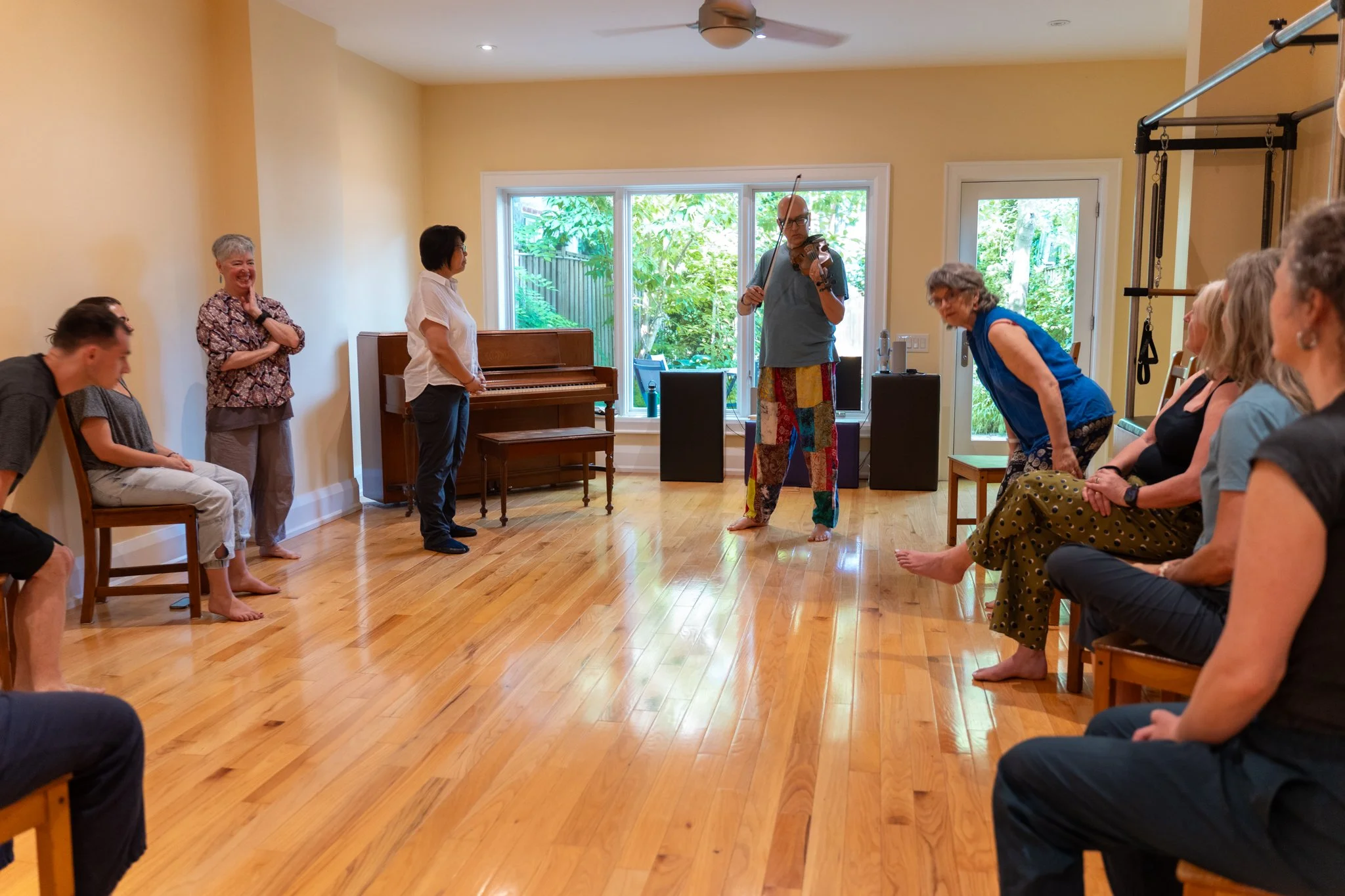 Group of people gathered in a living room watching a man with a violin perform, some seated and some standing, with a piano and large glass doors in the background.
