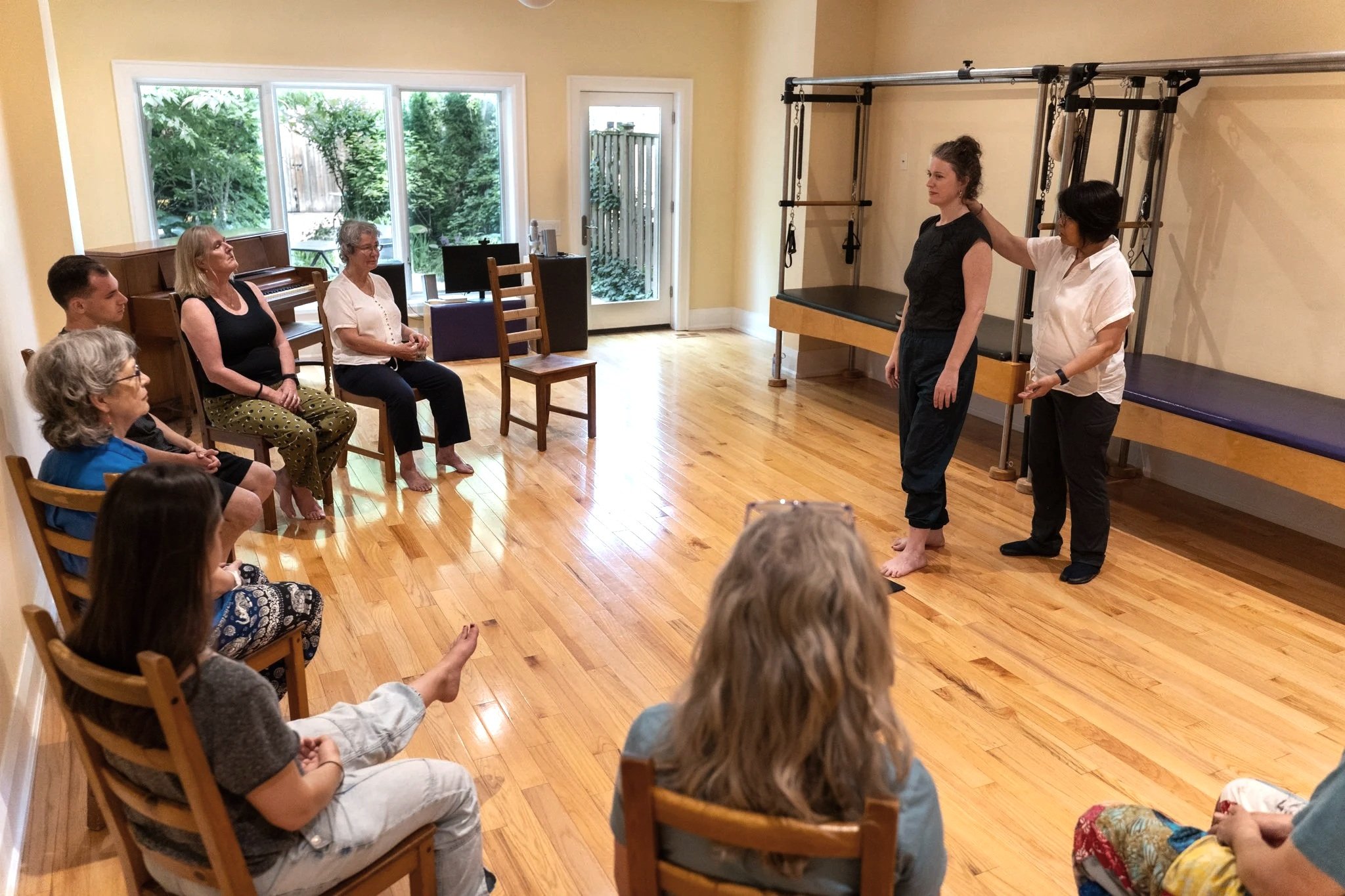Group of women sitting in a circle in a room with wooden floors, listening to two women at the front demonstrating movements or exercises. The room has large windows with greenery outside.