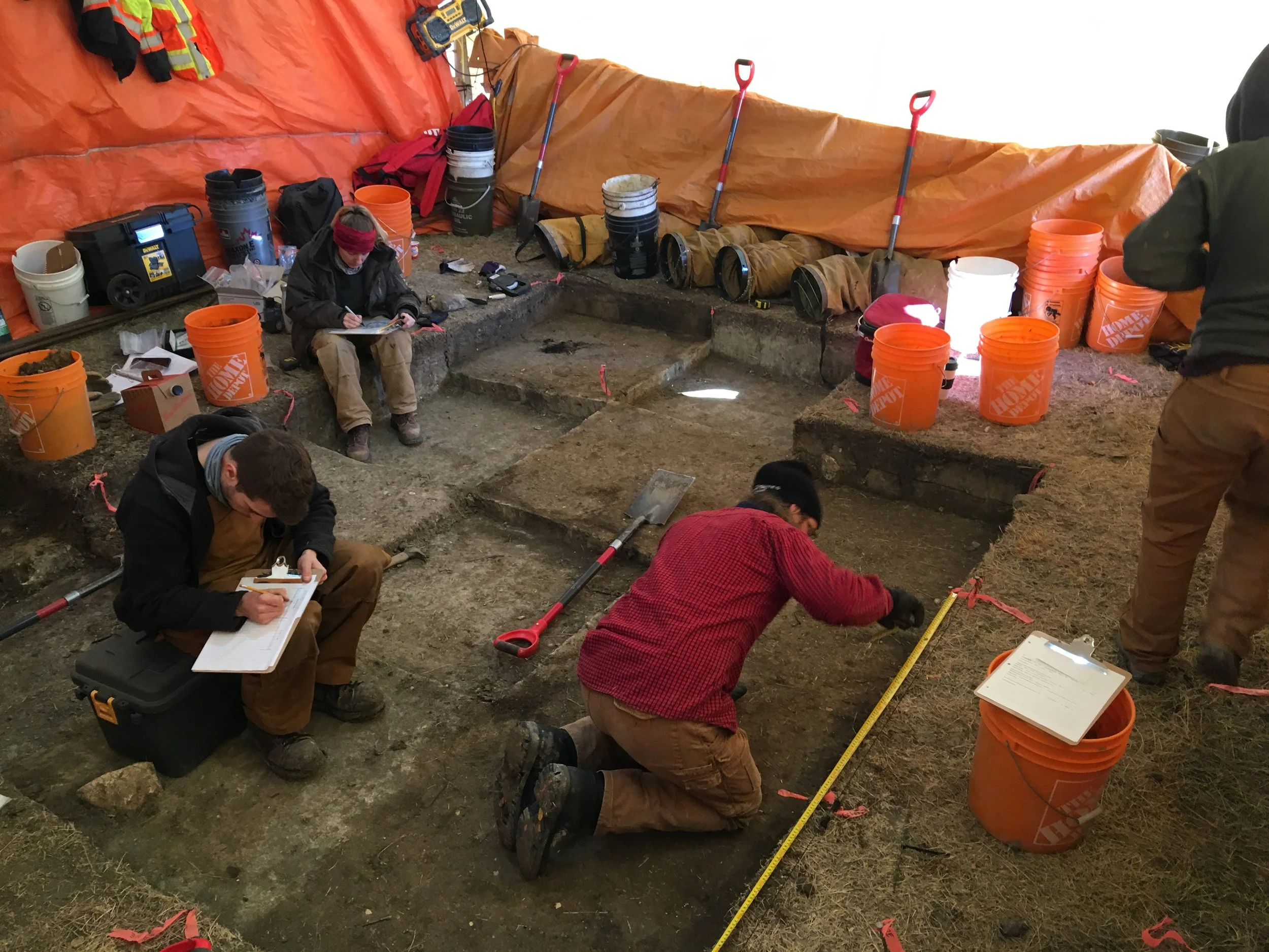 Archaeologists completing a field mitigation. Surrounded by field equipment and safety supplies. Measuring, digging, and assessing the field for potential artifacts.