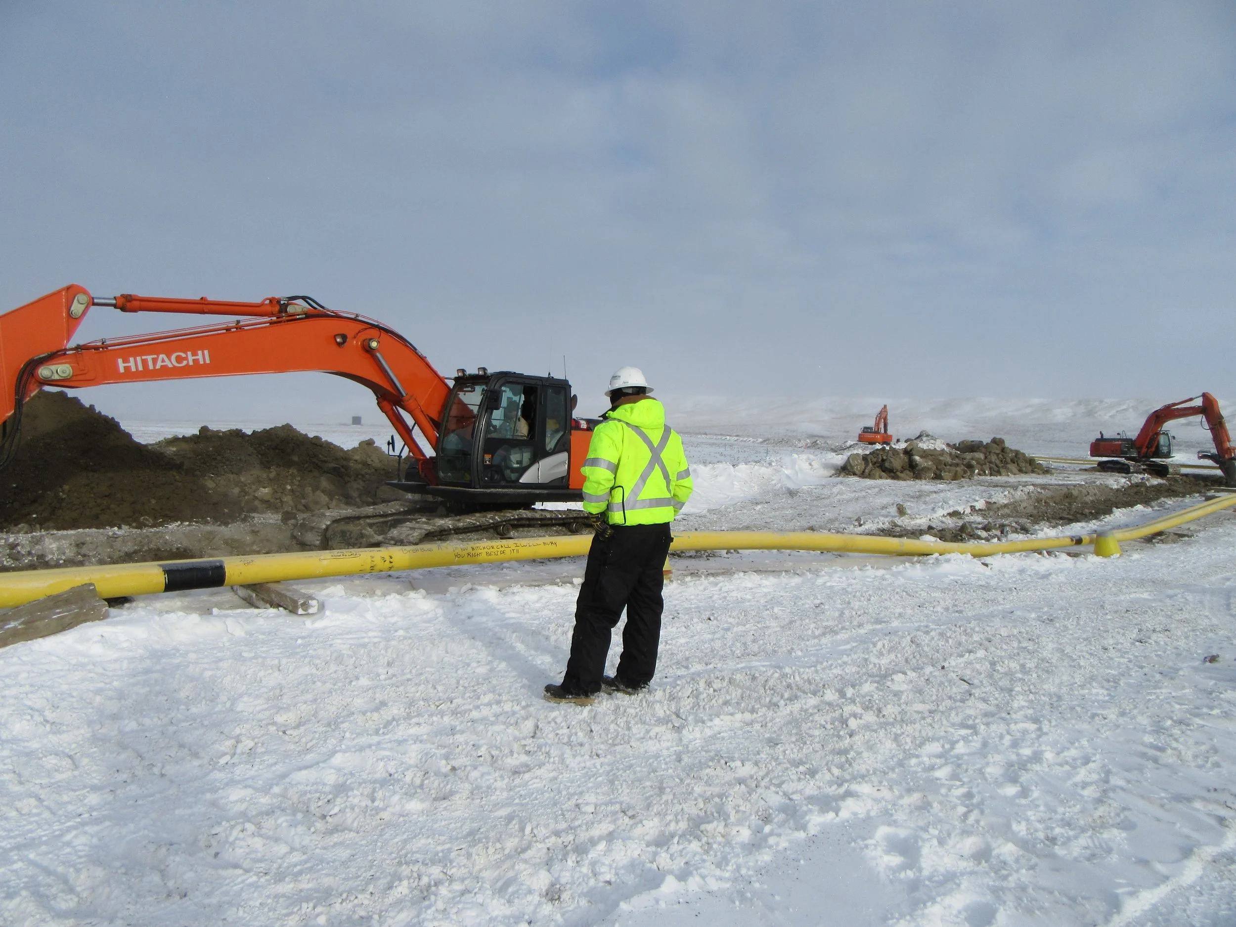 Archaeologist completing a heritage resources impact assessment in winter conditions. Professional is monitoring a site for potential sites with heavy equipment infront of them and snow covered ground landscape in Saskatchewan.