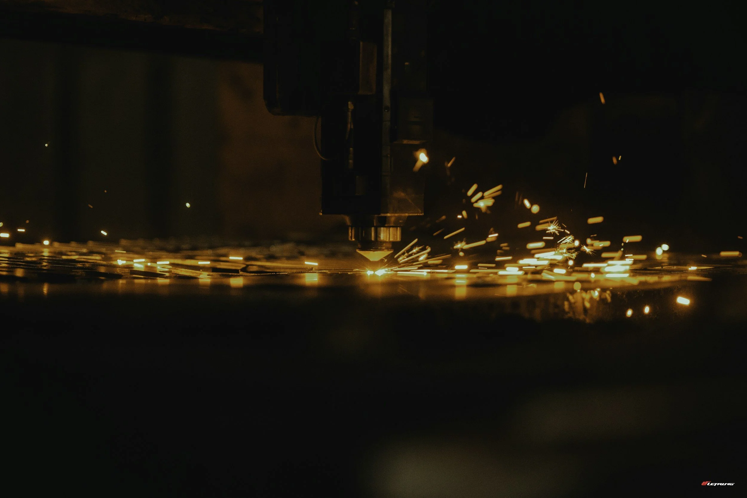 A close-up photo of a laser cutter in operation, emitting bright sparks and light as it cuts through metal.