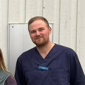 Mann i blå sykepleieruniform står foran en hvitvegget vegg