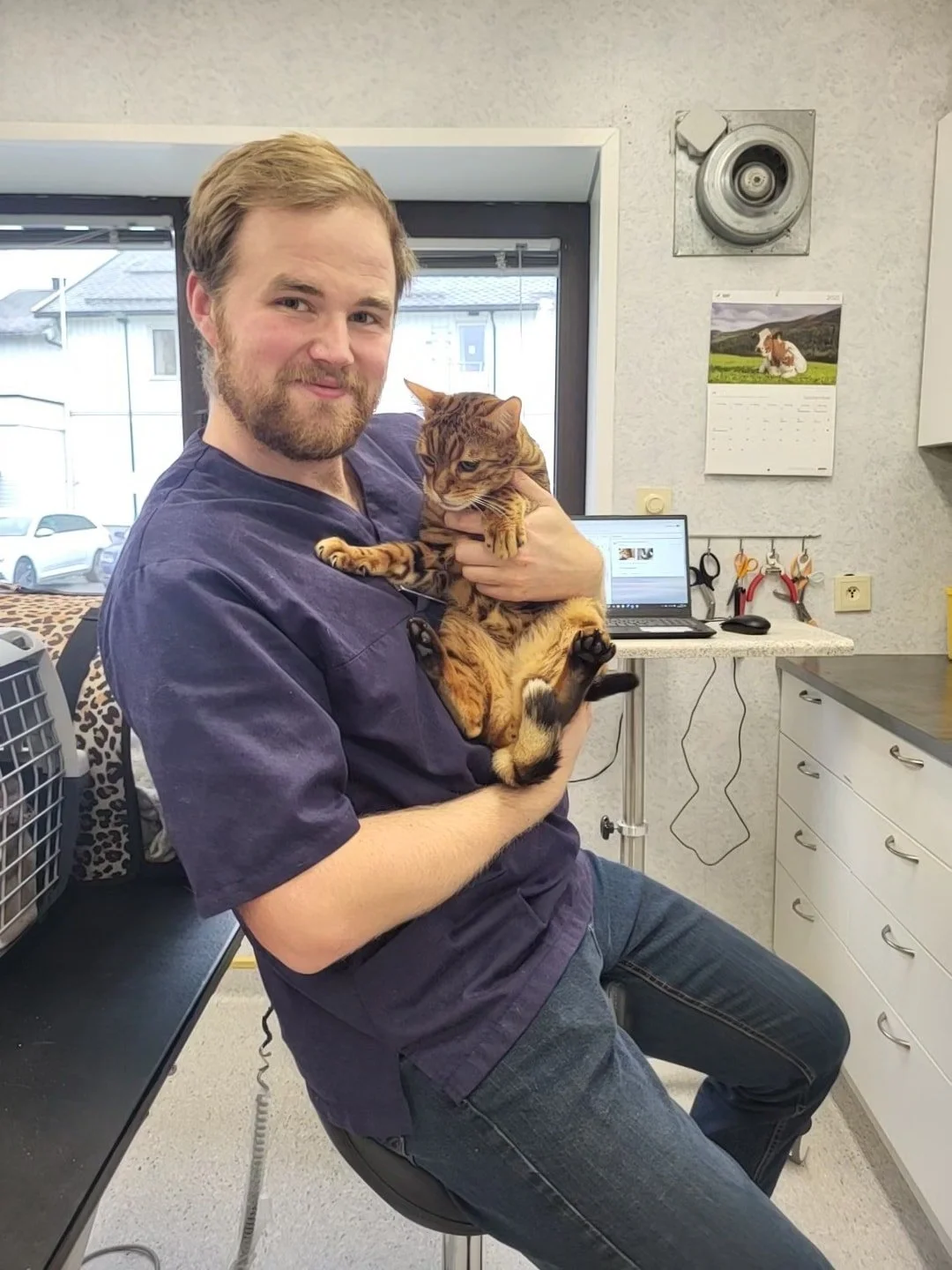 En mann sitter på en kontorstol med en katt i armene inne på et kontor eller klinikk, med datamaskin, verktøy og utstyr i bakgrunnen.