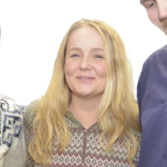 Et smilende kvinnemenneske med langt blond hår iført en strikket genser med et mønster.