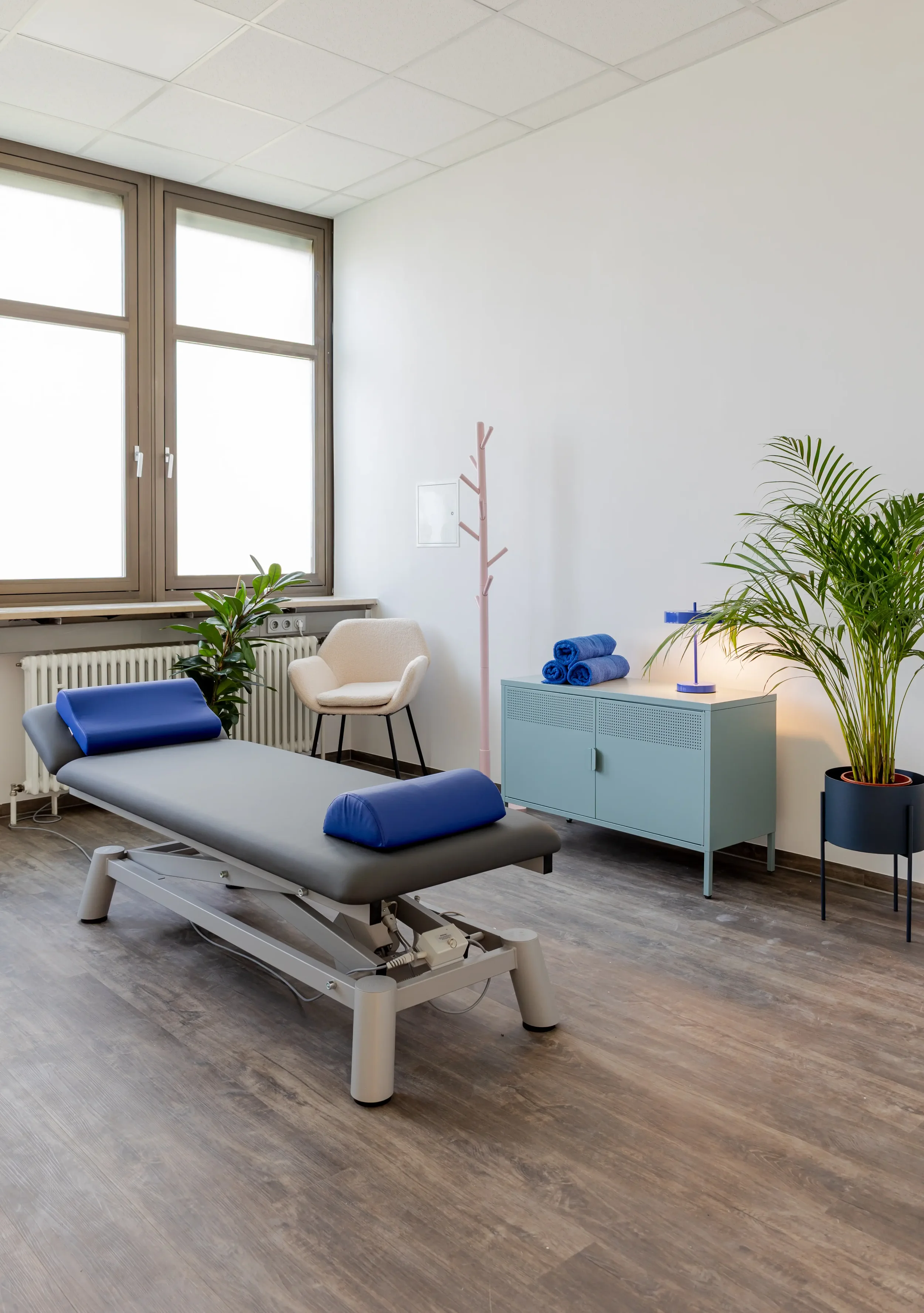 Therapieraum mit Behandlungsliege, Armlehnen und Kissen, Stuhl, Fenster, Pflanzen, Schrank mit Handtüchern und Lampe.