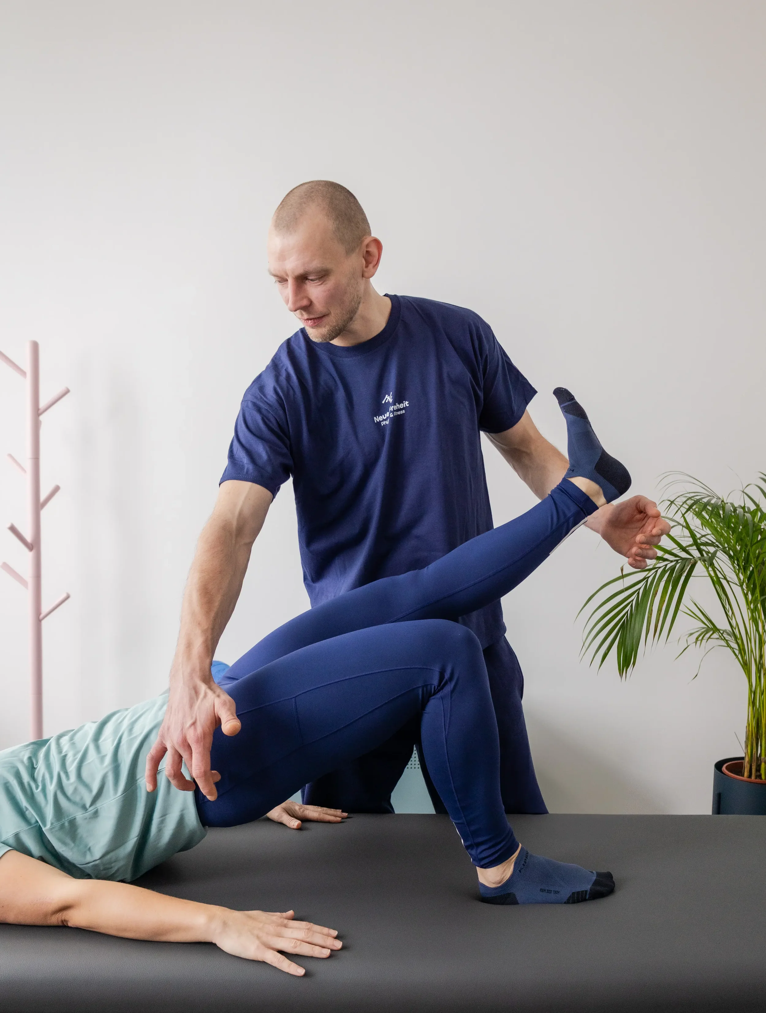 Ein Therapeut hält ein Bein eines Patienten bei einer Physiotherapie oder Fitnessübung auf einer Behandlungsliege. Im Hintergrund befindet sich eine Pflanze und eine pinkfarbene Kleiderständer.