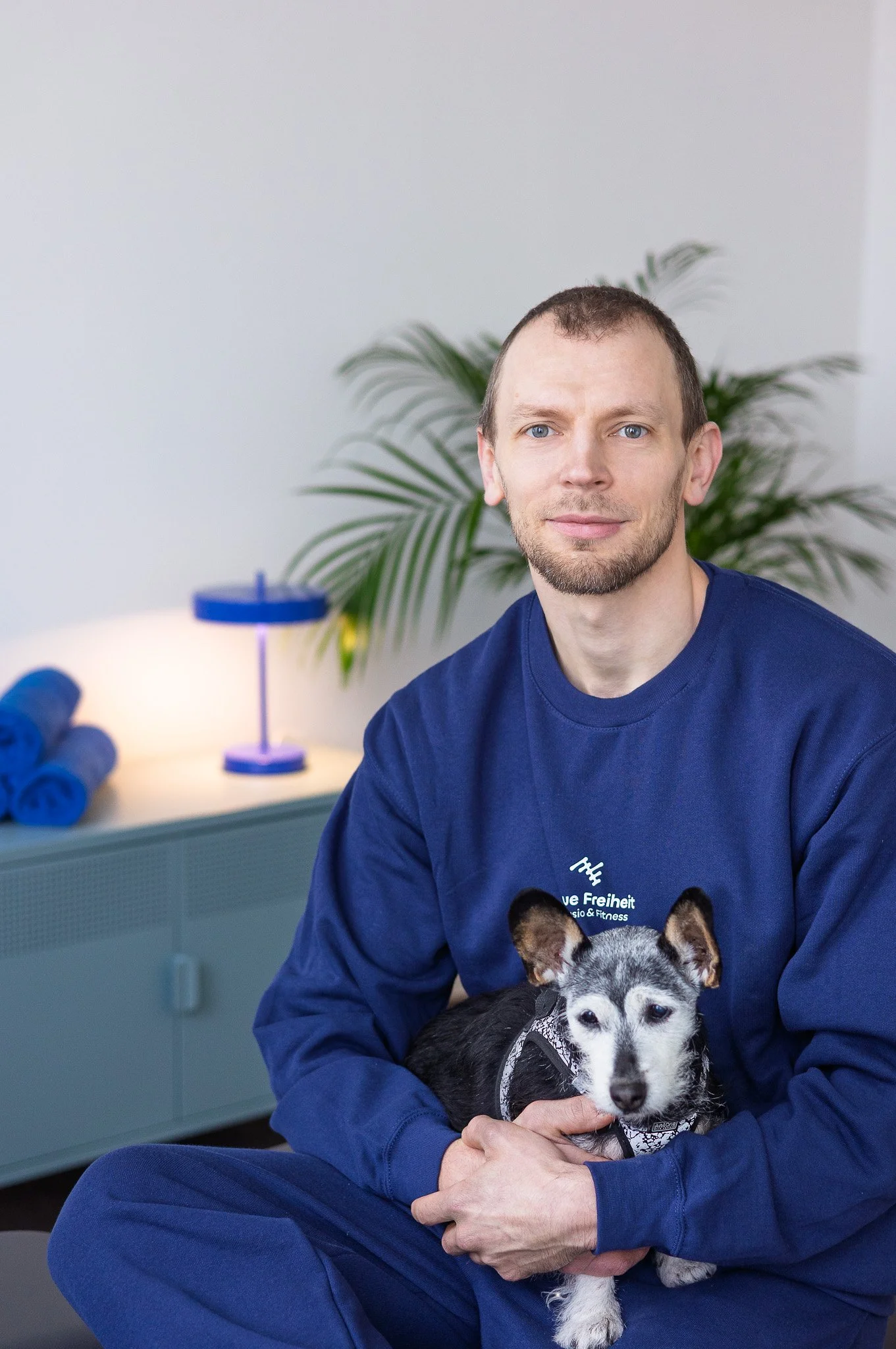 Ein Mann in blauer Sweatshirt hält einen kleinen Hund auf seinem Schoß in einem modern eingerichteten Raum mit grüner Pflanze und blauer Beleuchtung im Hintergrund.