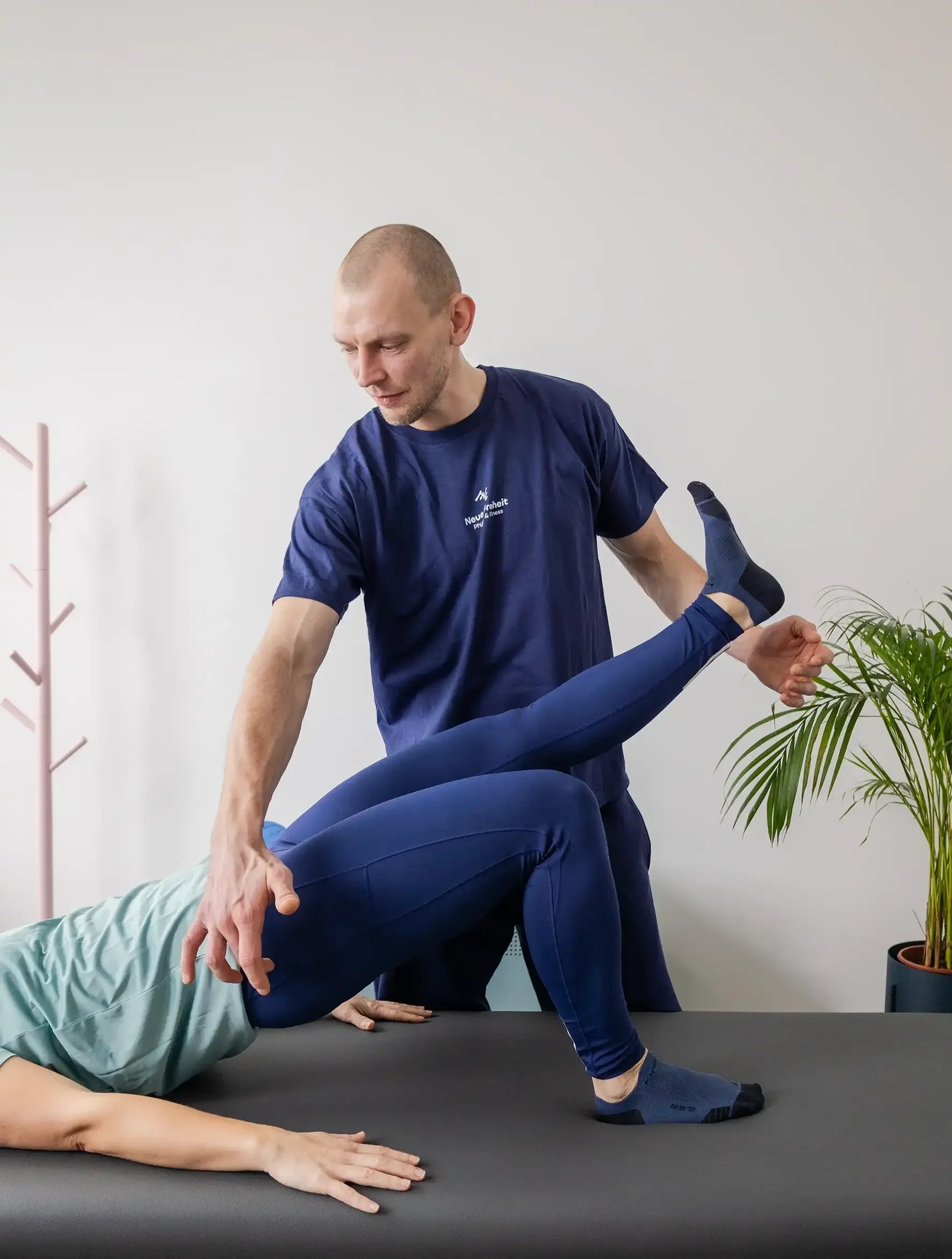 Ein Therapeut hält ein Bein einer Patientin bei einer Physiotherapie oder Fitnessübung auf einer Behandlungsliege. Im Hintergrund befindet sich eine Pflanze und ein pinkfarbener Kleiderständer.