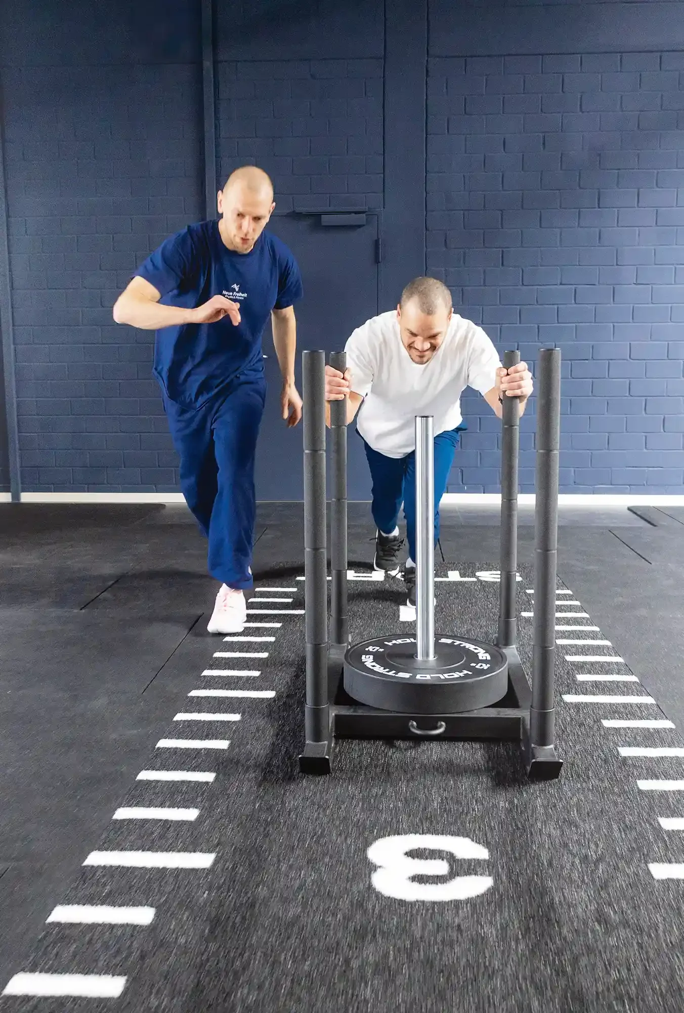 Ein Physiotherapeut von Neue Freiheit Physio & Fitness macht eine Übung mit einem Patienten. Der Patient schiebt einen Schlitten, der Therapeut begleitet ihn.