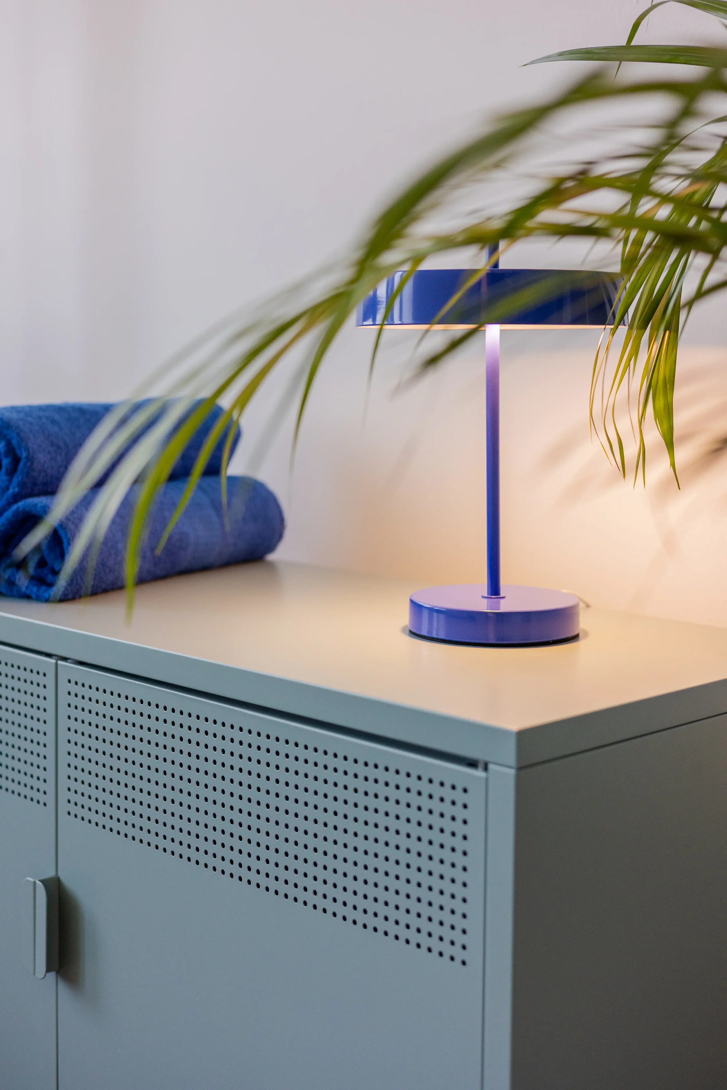 Ein blauer Tischlampenleuchte steht auf einem weißen Sideboard, daneben liegt ein zusammengerolltes blaues Handtuch. Im Vordergrund befinden sich grüne Pflanzenblätter.