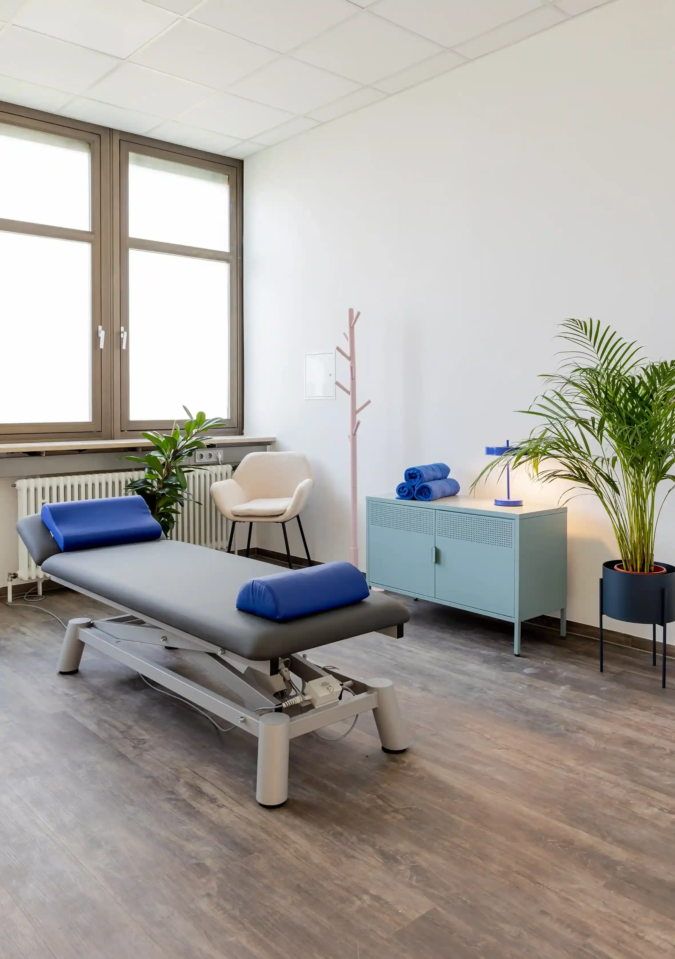 Ein Behandlungsraum einer Physiotherapie. Im Vordergrund steht eine Therapieliege. Im Hintergrund steht ein Stuhl und diverse Einrichtungsgegenstände. Warmes Tageslicht scheint durch die Fenster.