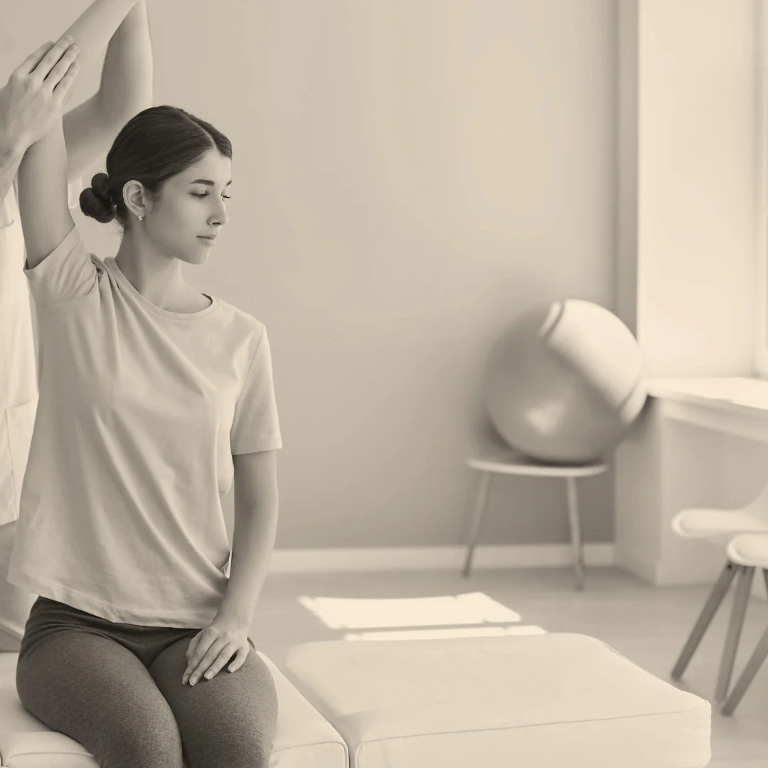 Woman receiving physiotherapy, sitting on a mat with a therapist assisting arm mobility exercises in a bright room with an exercise ball in the background.