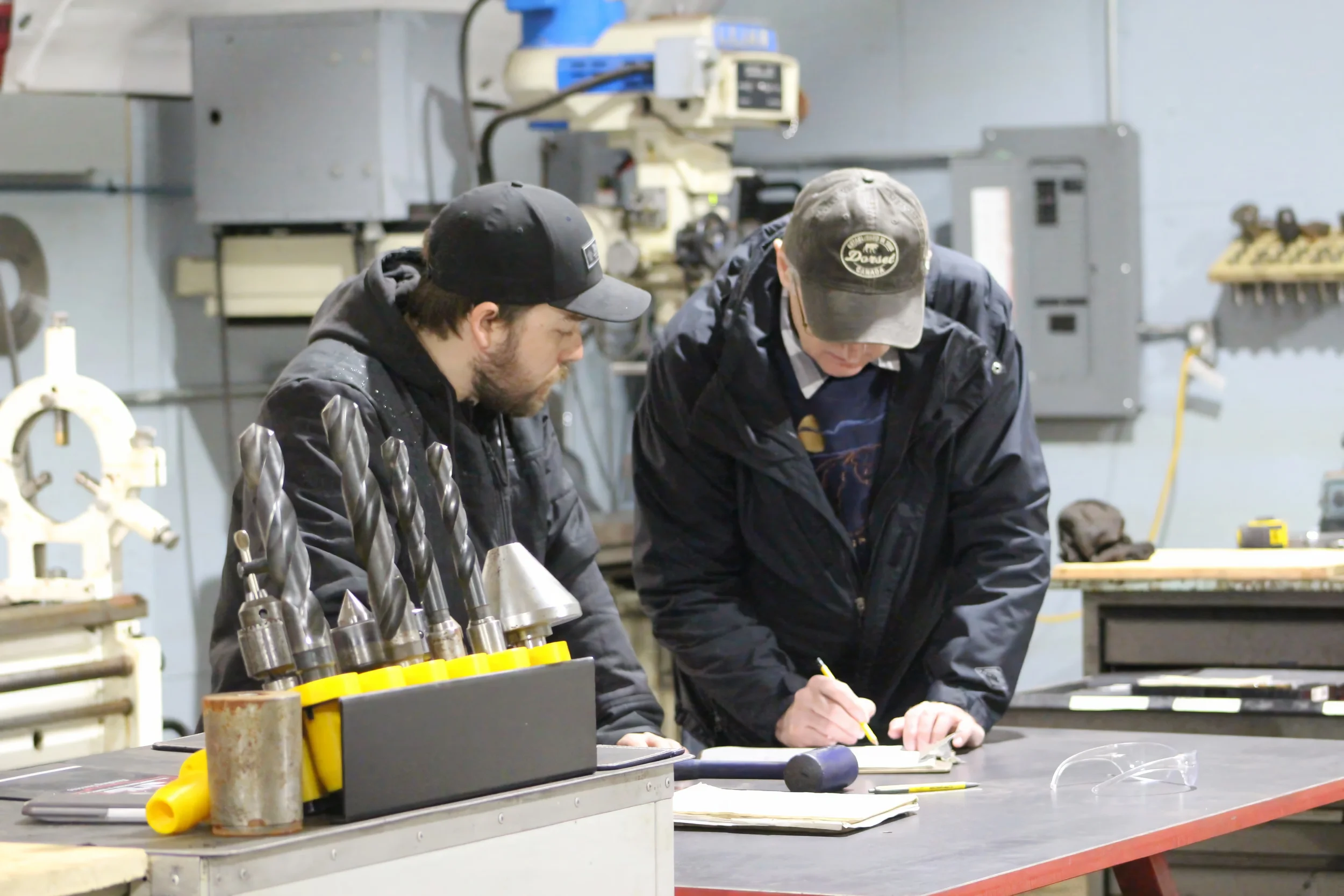 Two men working at a table in a workshop, with various tools and equipment around them, focused on a project.