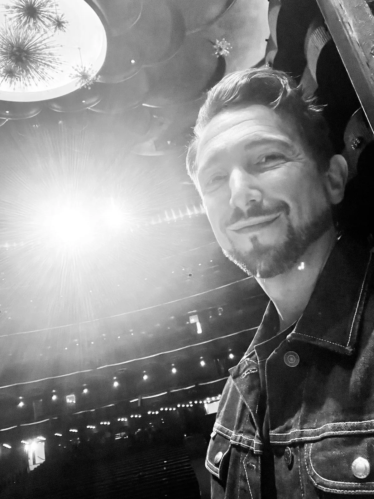 A black and white shot of Aaron Blake in a theater with stage lights and chandeliers overhead.