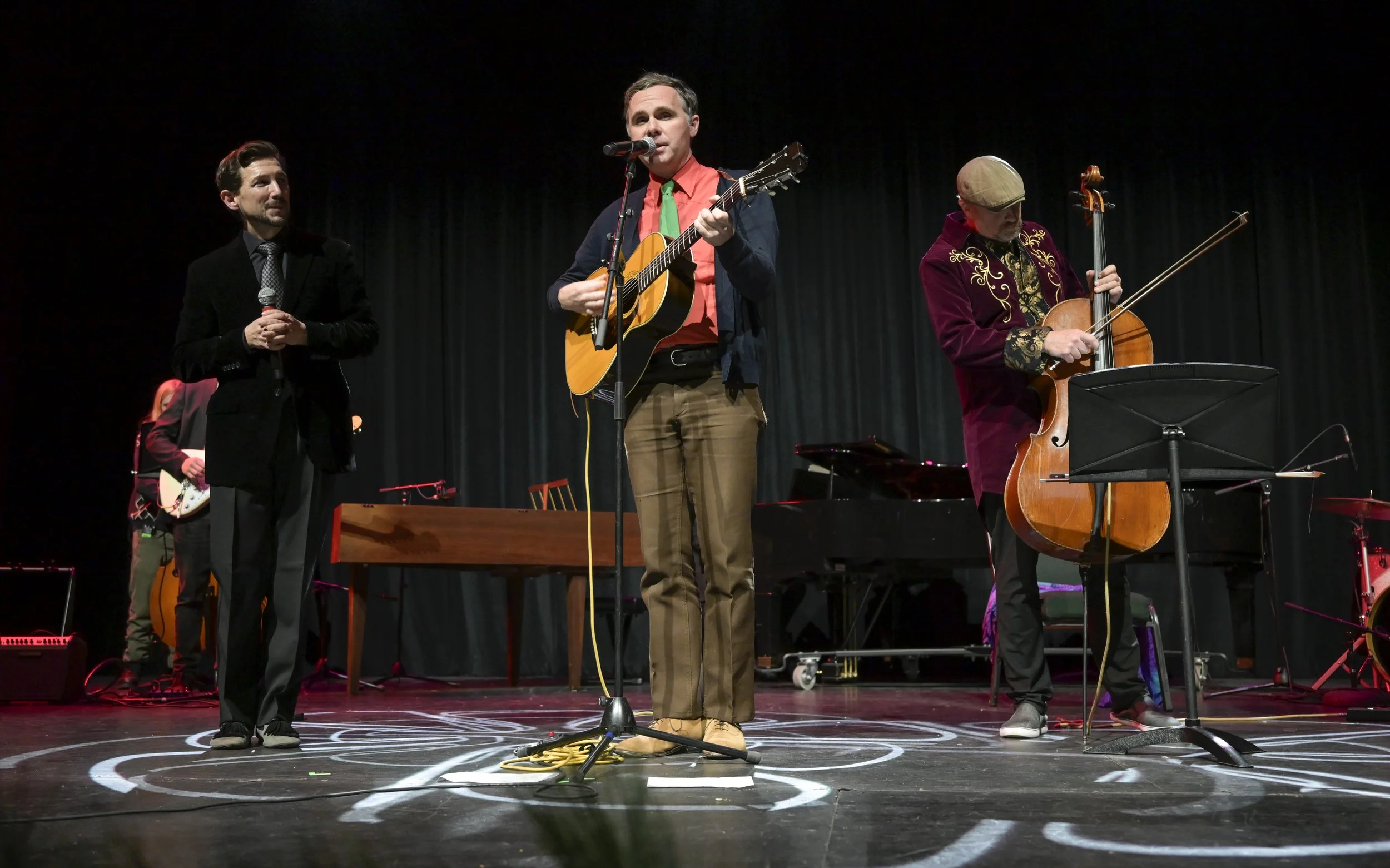 Kris Truelsen, Aaron Blake, and Dave Eggar performing on stage with instruments including a guitar, cello, and piano, in front of black curtains.