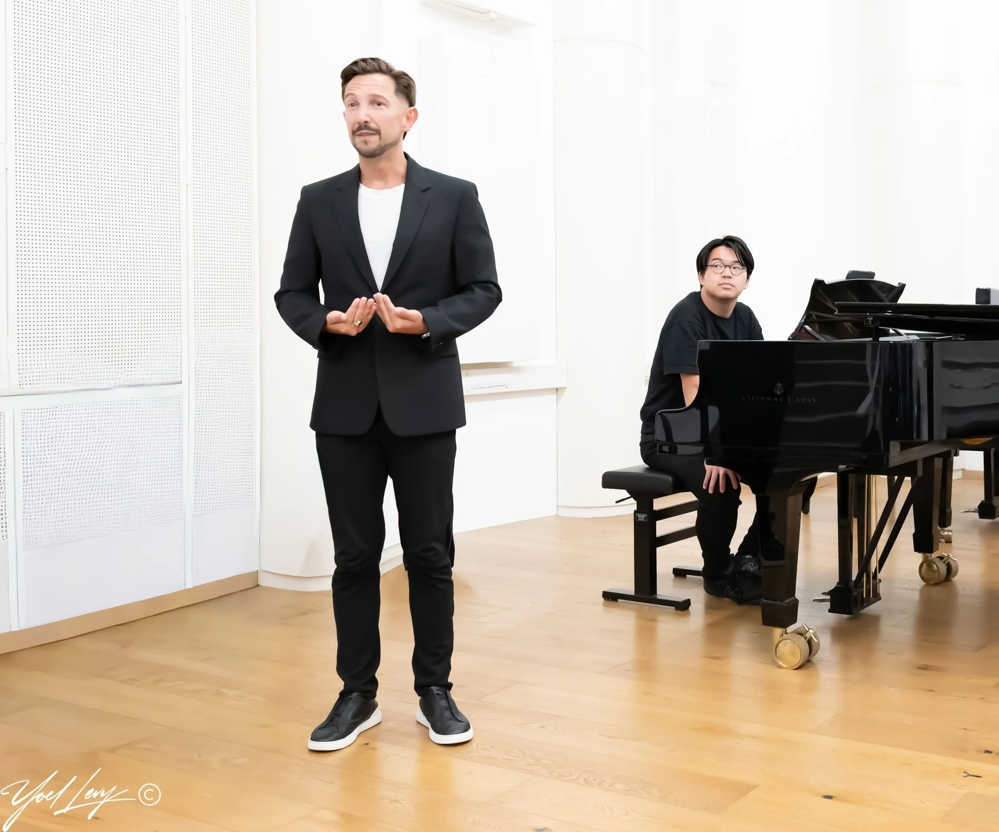 A man in a black suit stands in front of a piano in a music studio