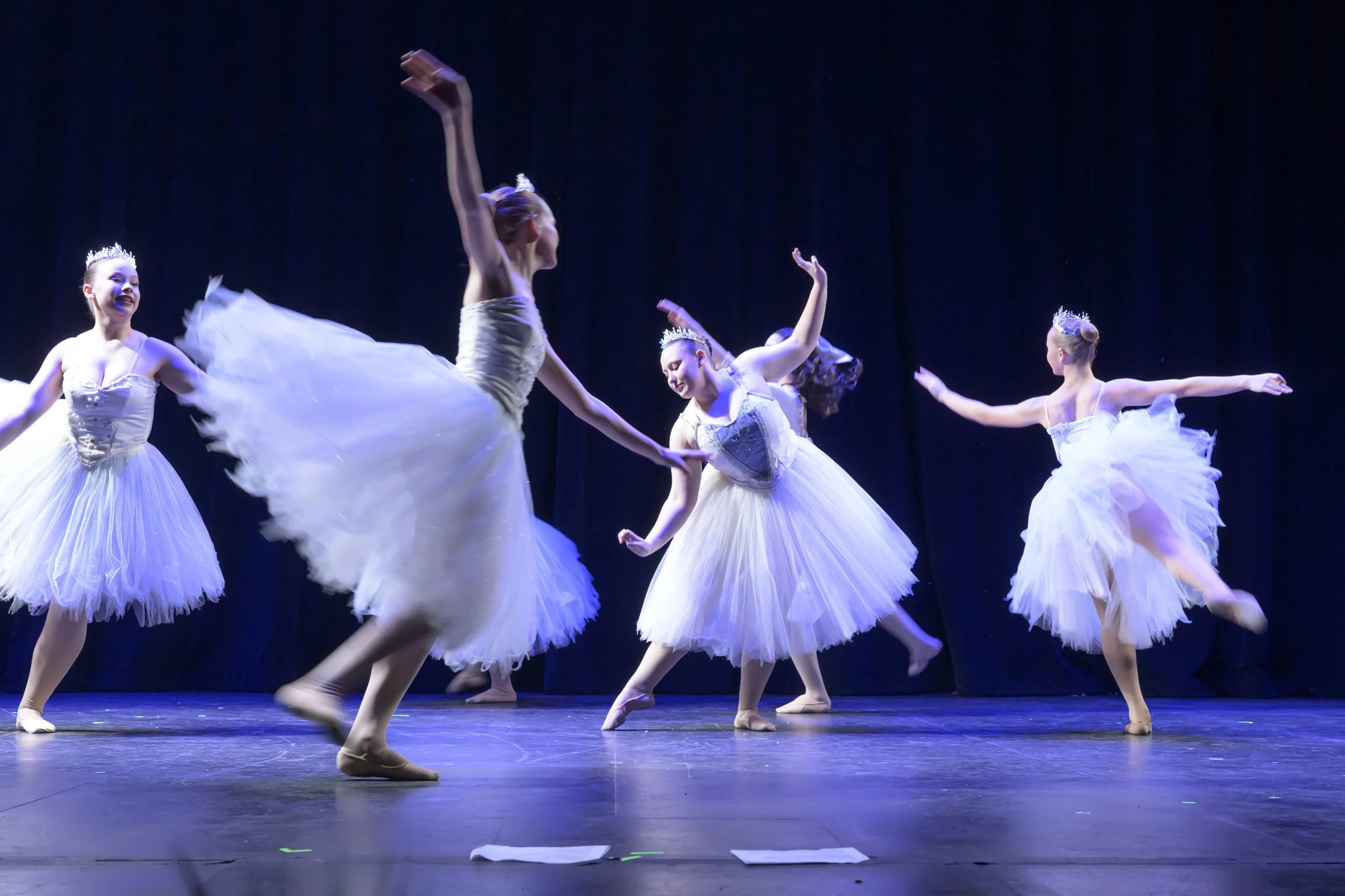 Ballet dancers in tutus performing on stage with dark background, some with tiaras, in graceful poses.