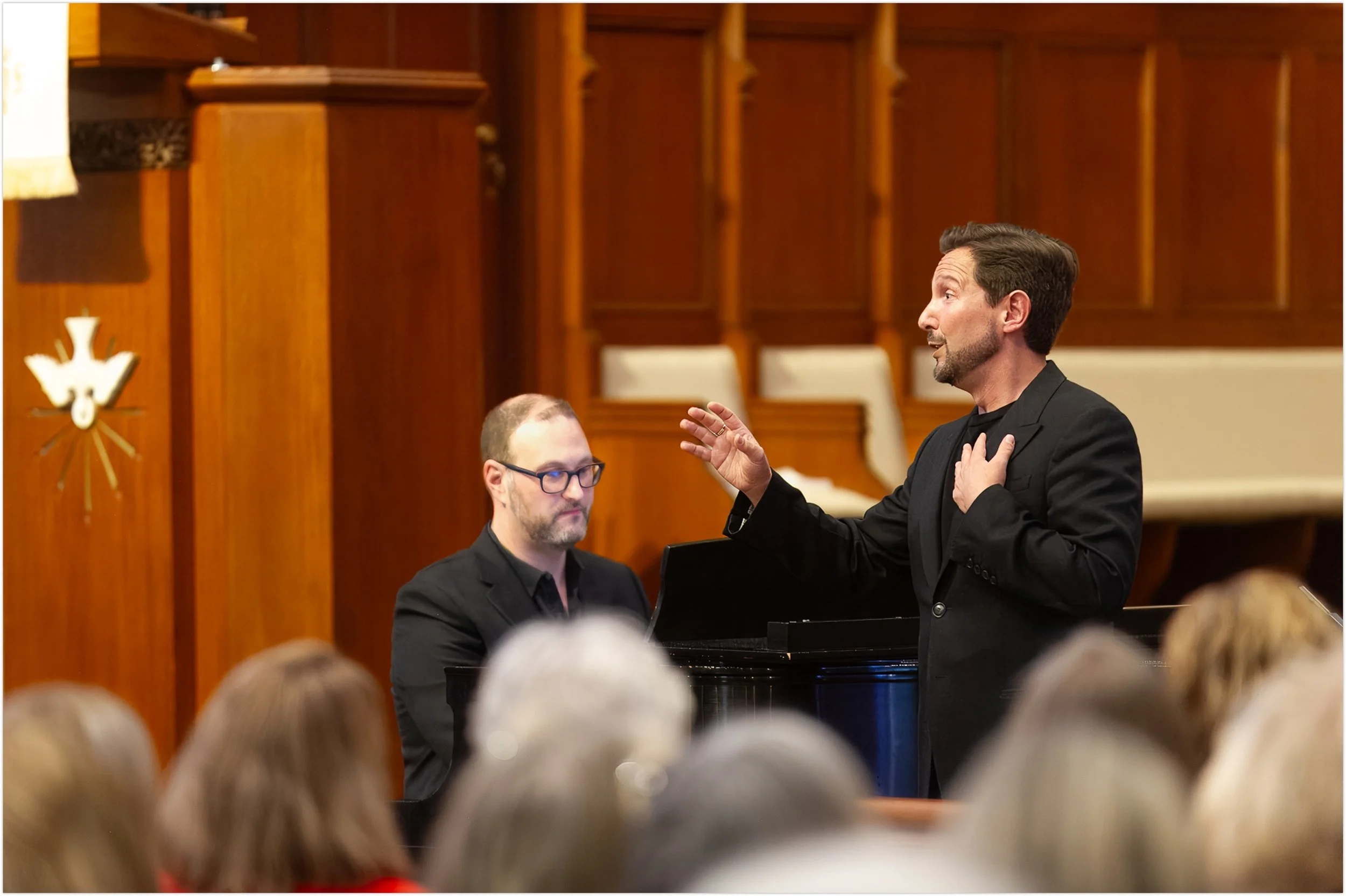 Grammy-winning tenor Aaron Blake in a black suit singing with his hand on his chest, standing next to a seated man at a piano. The scene takes place in a wood-paneled sanctuary with an audience in the foreground.