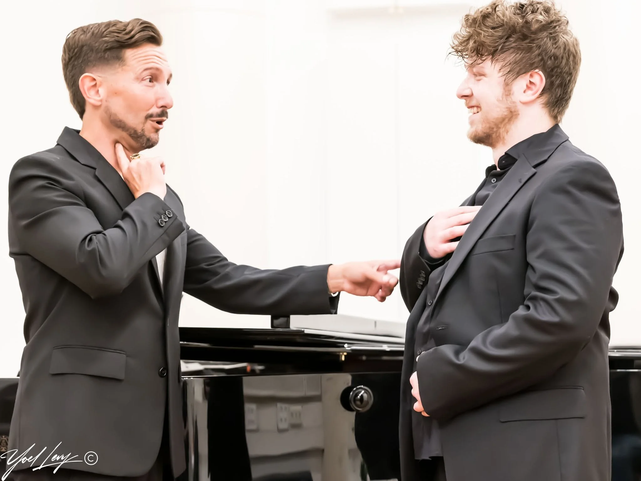 A bearded man in a dark suit teaches a vocal arts student in front of a piano