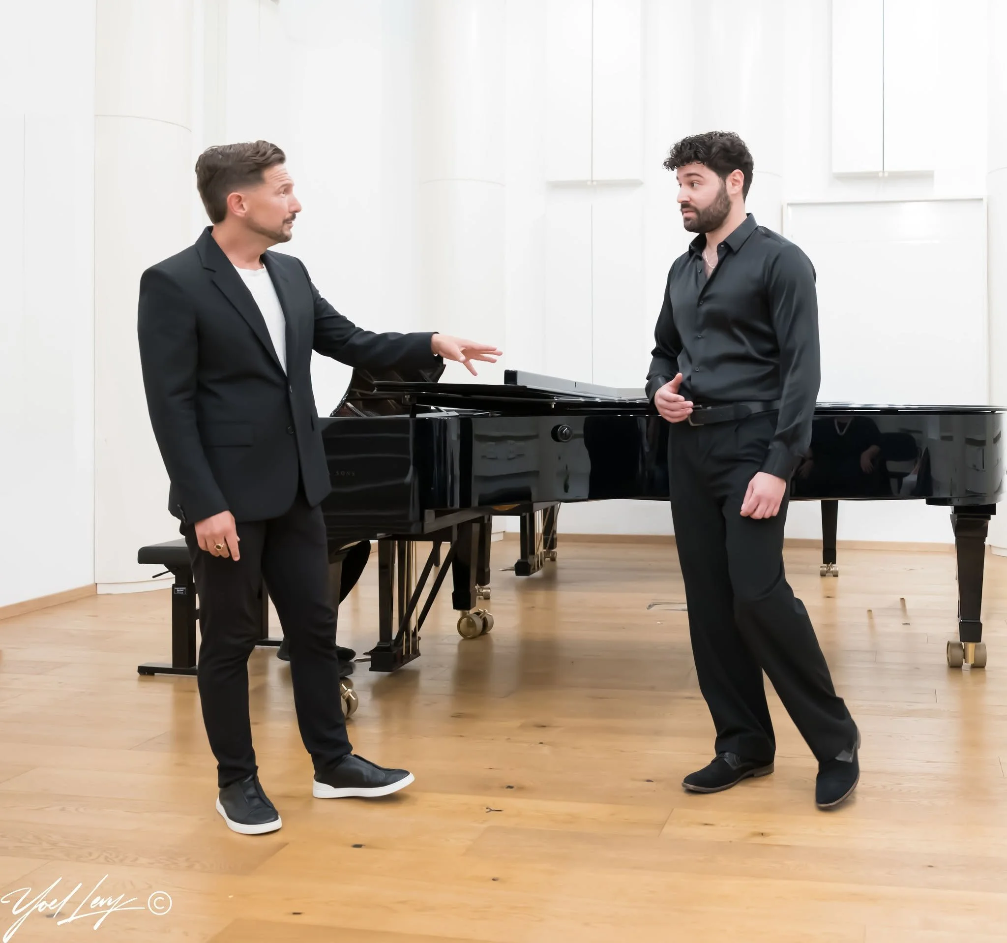 Two men in dark suits stand in front of a piano