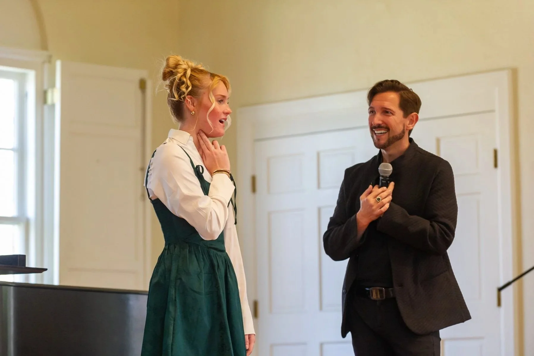 Aaron Blake in a black suit, holding a microphone and smiling, talking to a woman with blond hair, dressed in a dark green dress and white blouse. They are indoors with a cream-colored wall and white double doors in the background.