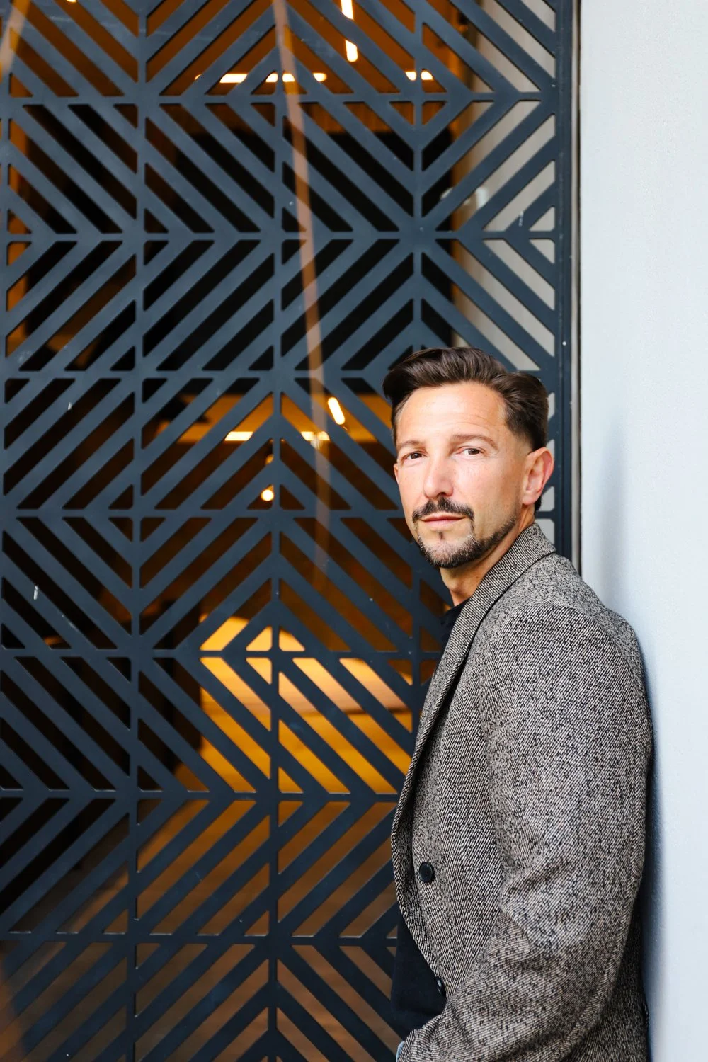 A bearded man leaning against a white wall with a geometric gate in the background