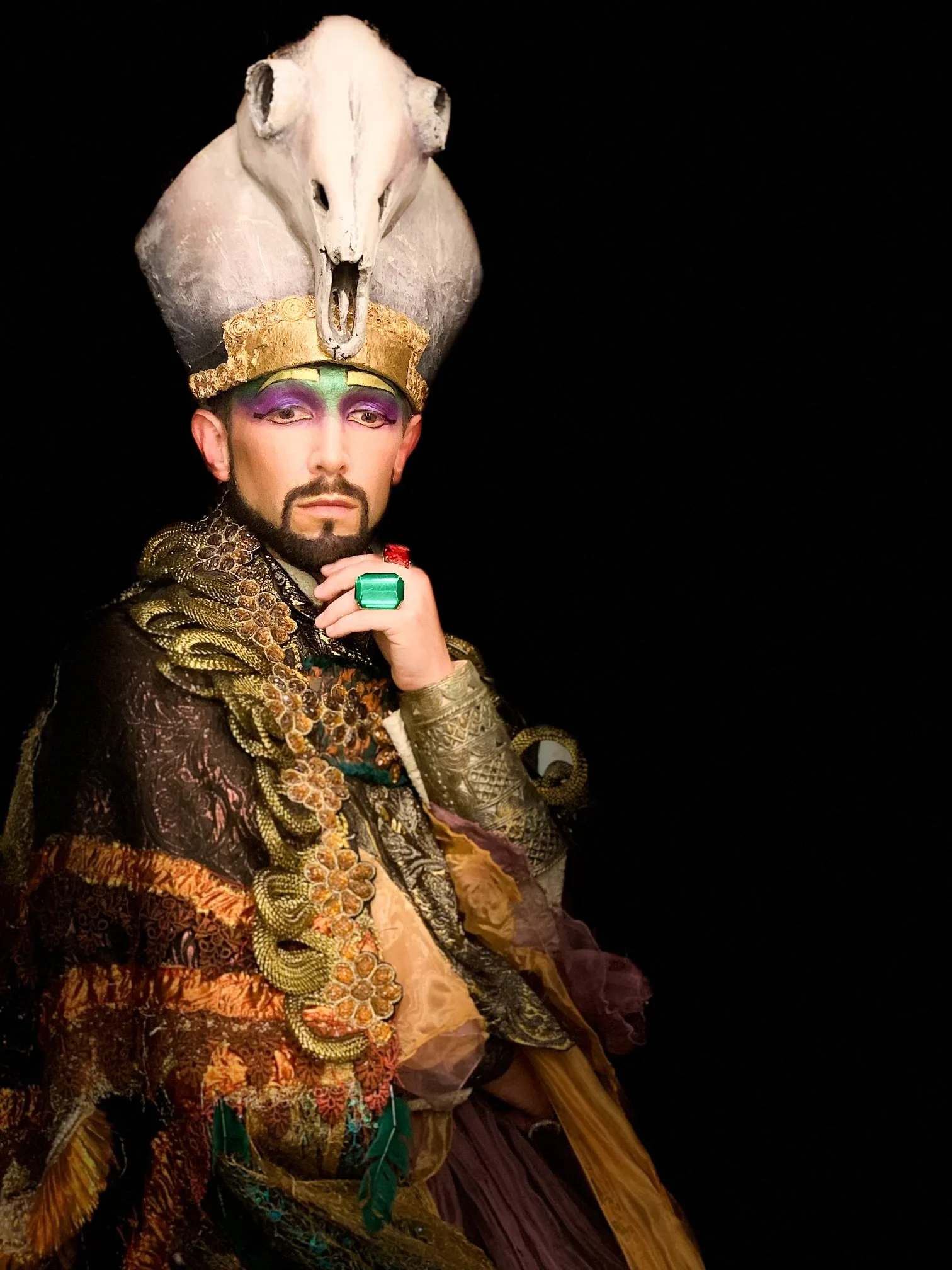 Aaron Blake in an elaborate Egyptian-style costume with ornate gold jewelry, wearing a headdress featuring an animal skull and bold makeup posing against a black background.