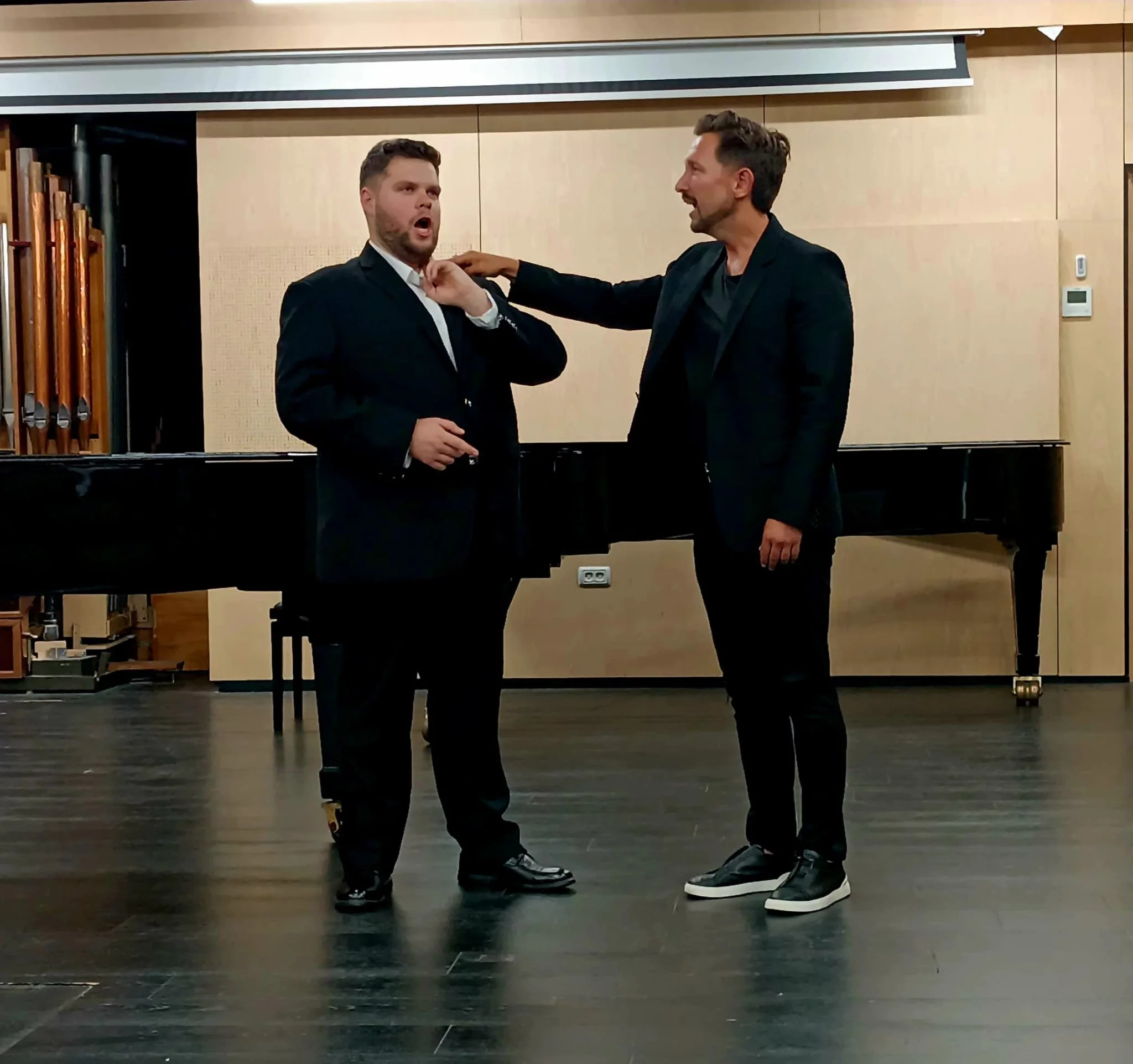 Two men in dark suits sing in front of a grand piano