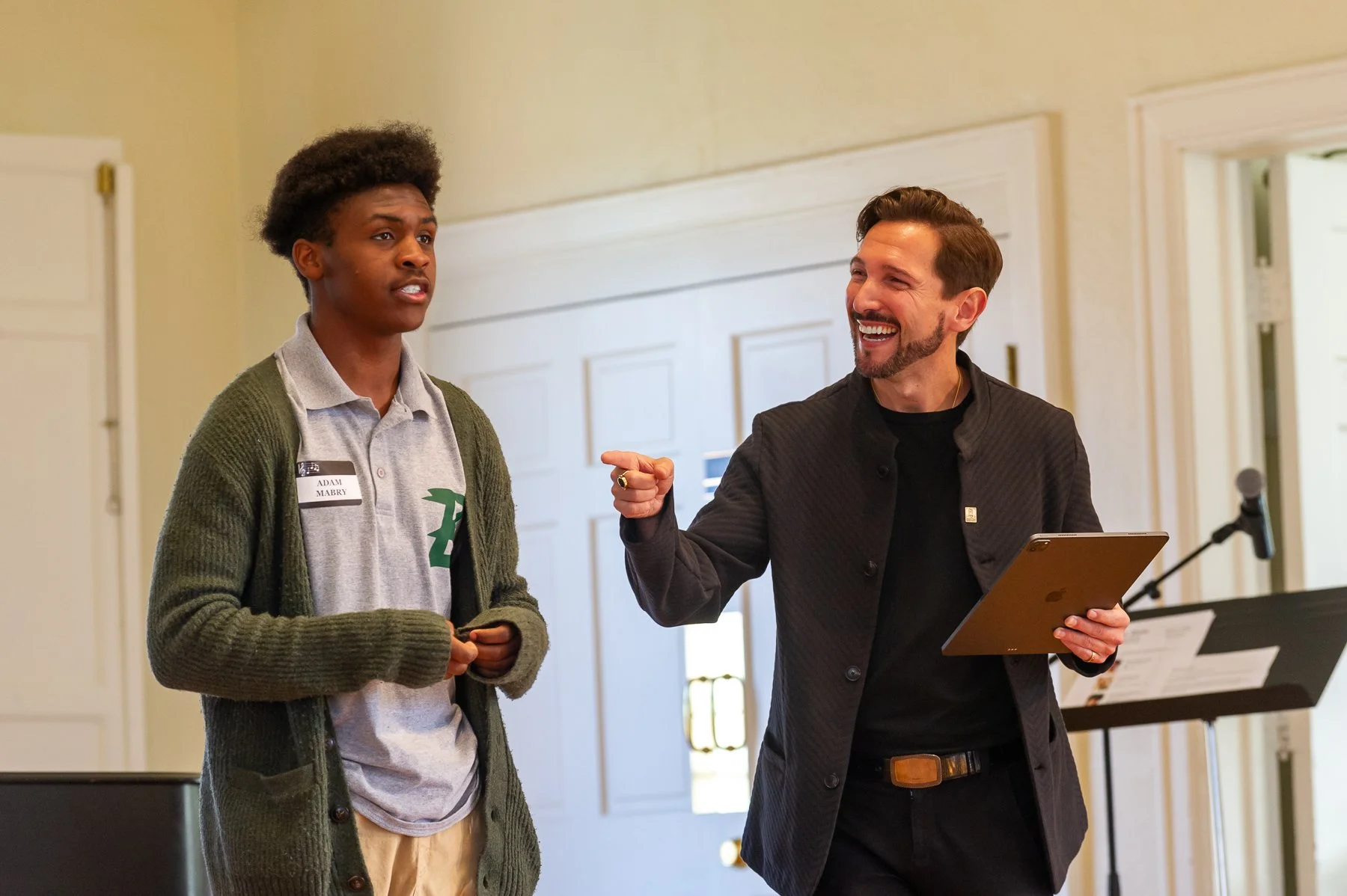 Aaron Blake smiling and pointing to a young man who is singing, with a lectern and microphone nearby.
